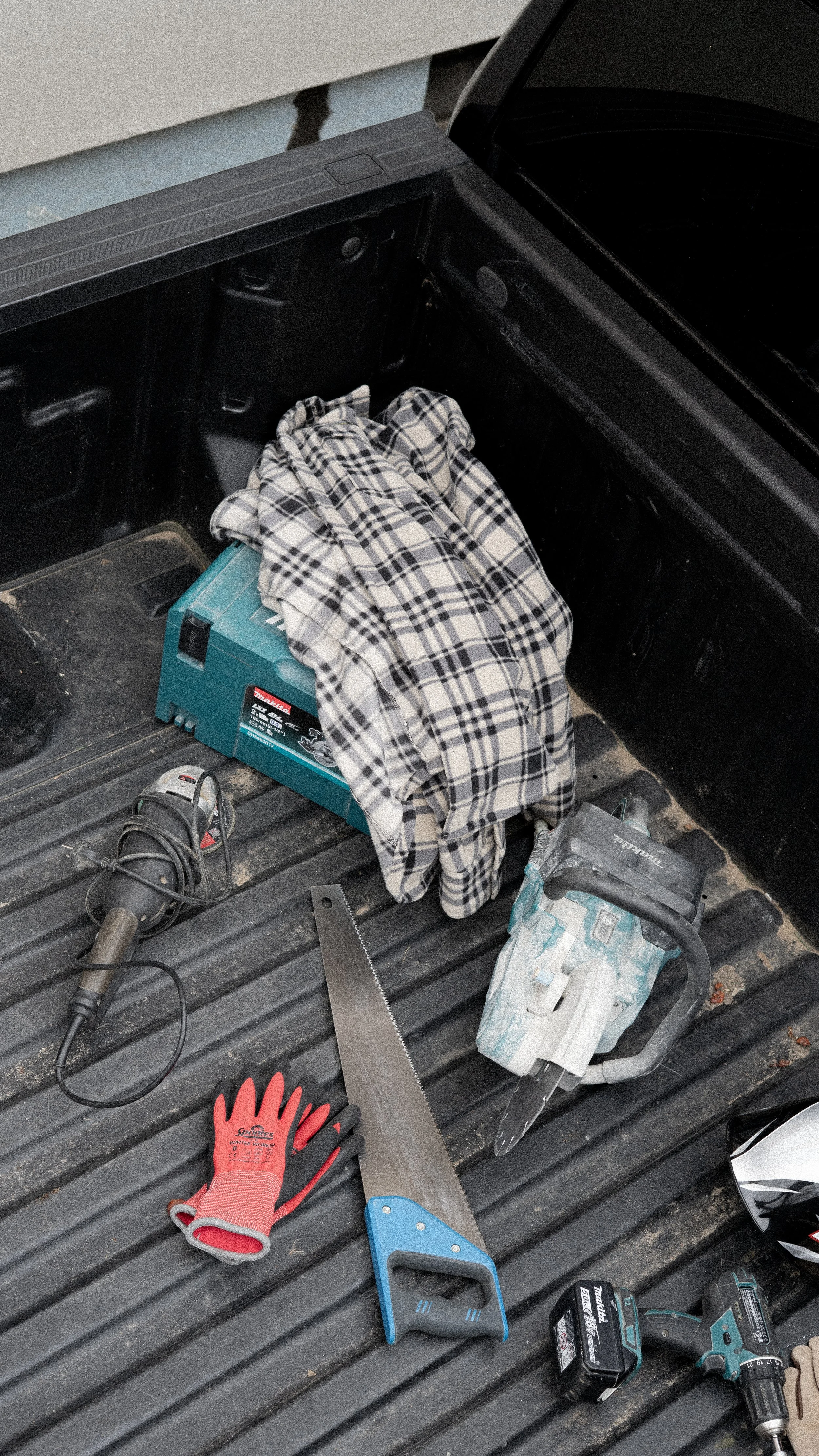 Werkzeug und Arbeitsausrüstung im Kofferraum eines Autos, inklusive Schweißermaske, Säge, Akkuschrauber, Handschuhe, Karohemd, Kabel und Kleidung.