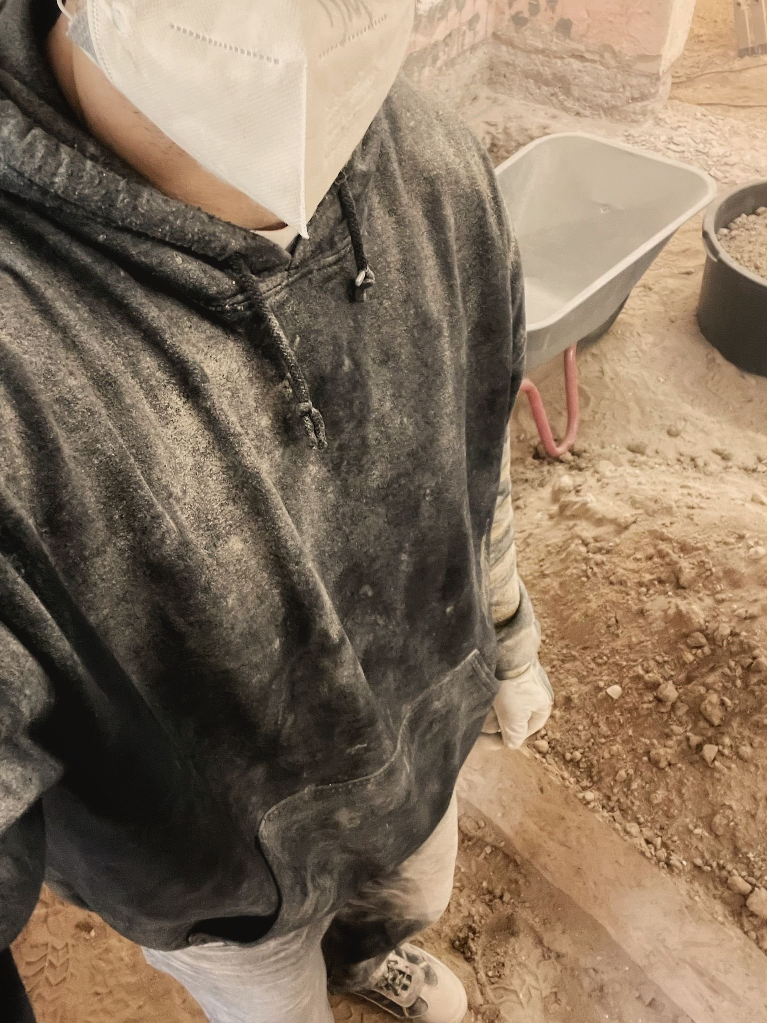 Person trägt verschmutzten schwarzen Hoodie und weiße Jogginghose, im Hintergrund Baustellenumgebung mit Sand und Baufahrzeugen.