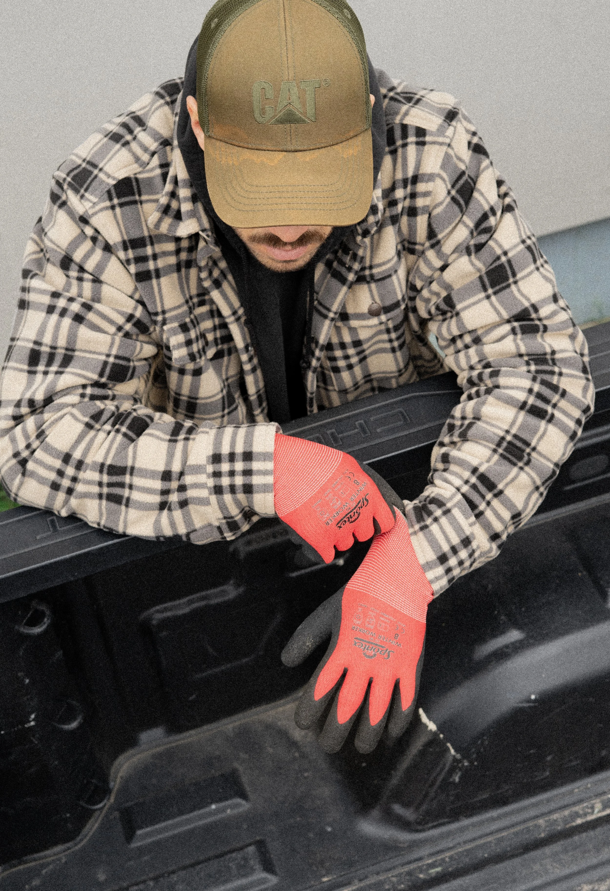 Ein Mann trägt eine beige Kappe mit dem Logo von CAT, ein kariertes Hemd und rote Arbeitshandschuhe, während er an einem schwarzen Fahrzeug arbeitet.