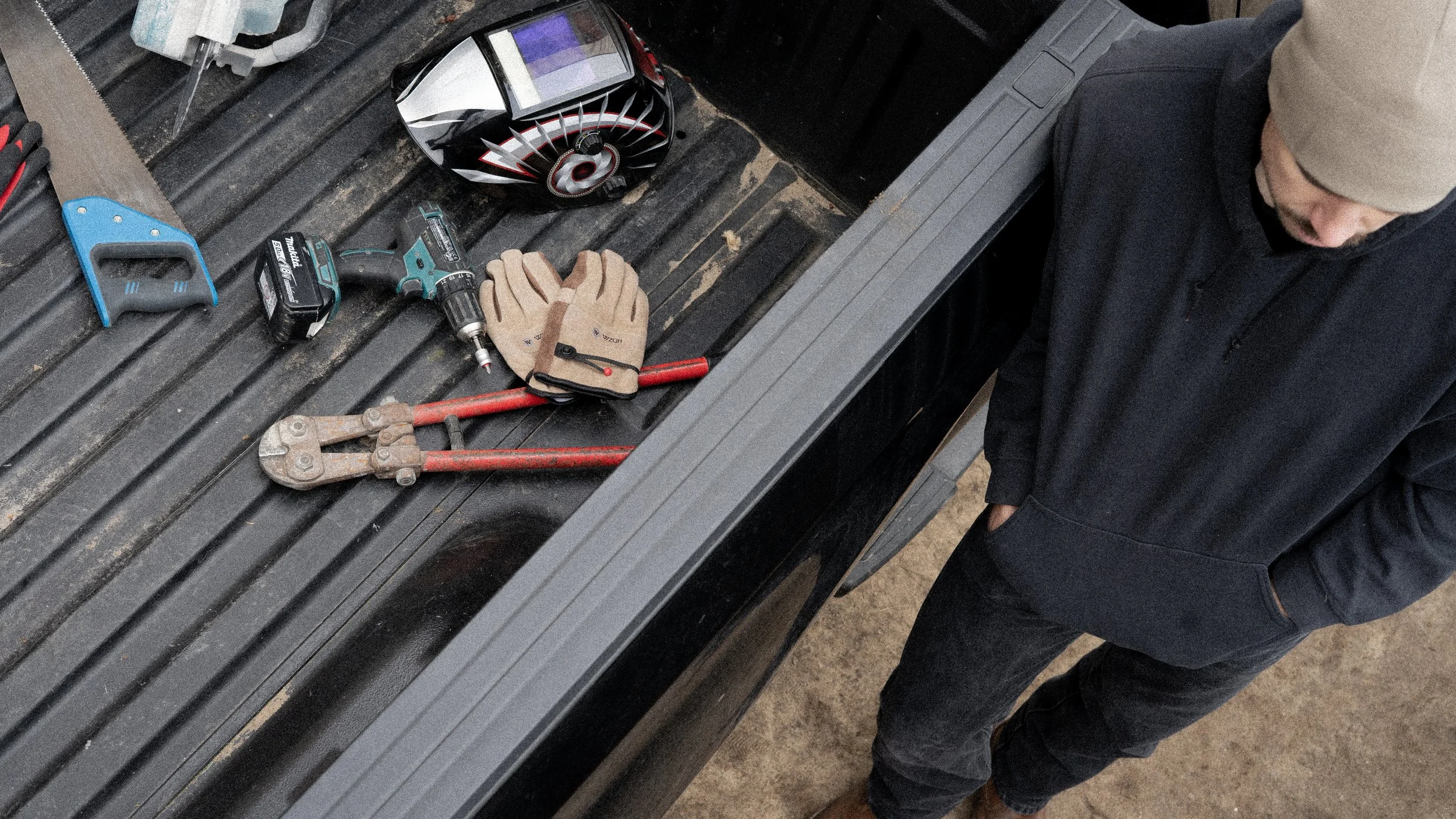 Werkzeug und Schutzbekleidung auf einem schwarzen Werkbank, daneben eine Person in dunkler Kleidung und Mütze, die Blick nach unten hat.