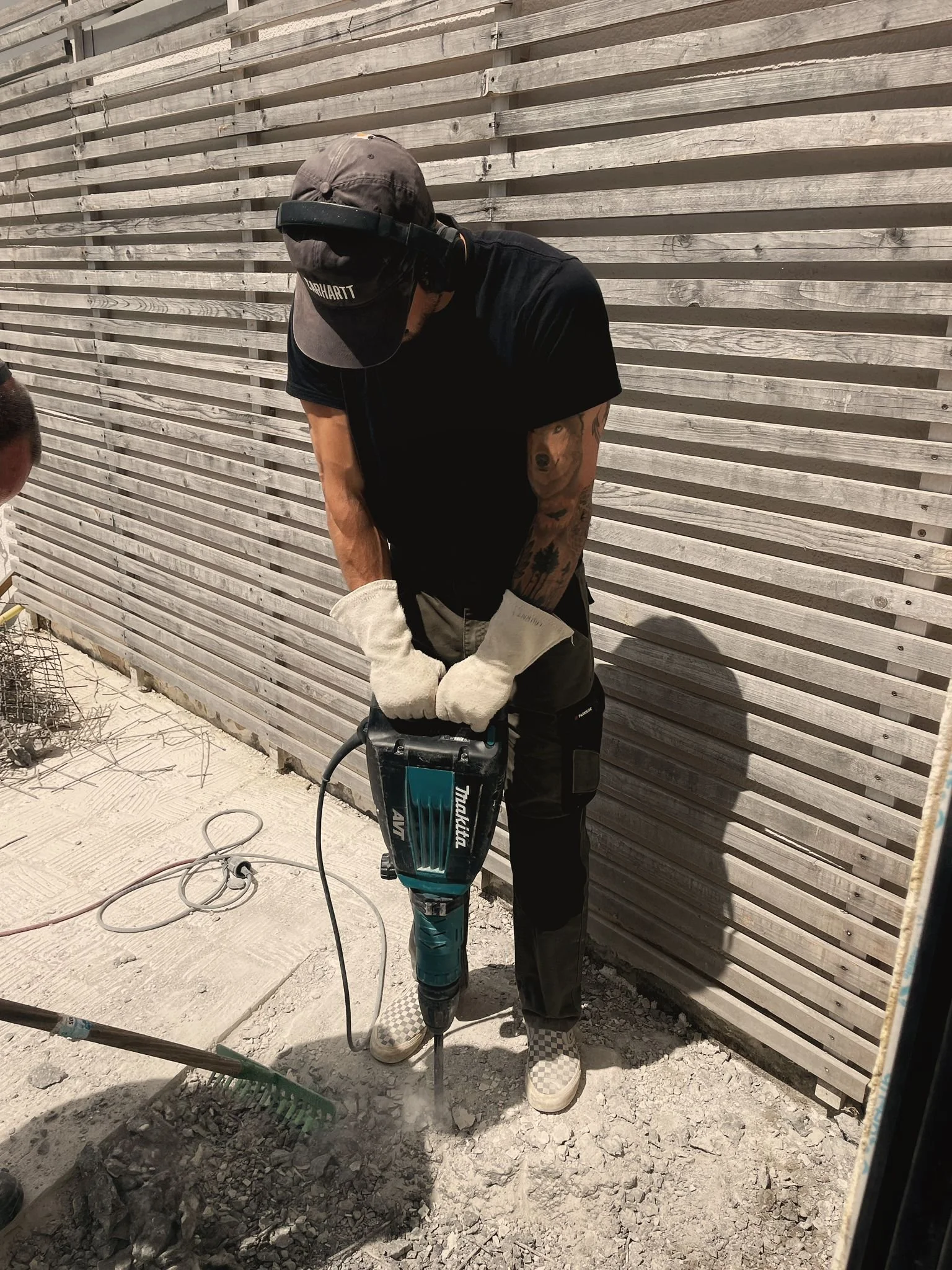 Mann benutzt einen Bohrer auf einem Boden, trägt Handschuhe und ein schwarzes T-Shirt, steht vor einer Holzwand.