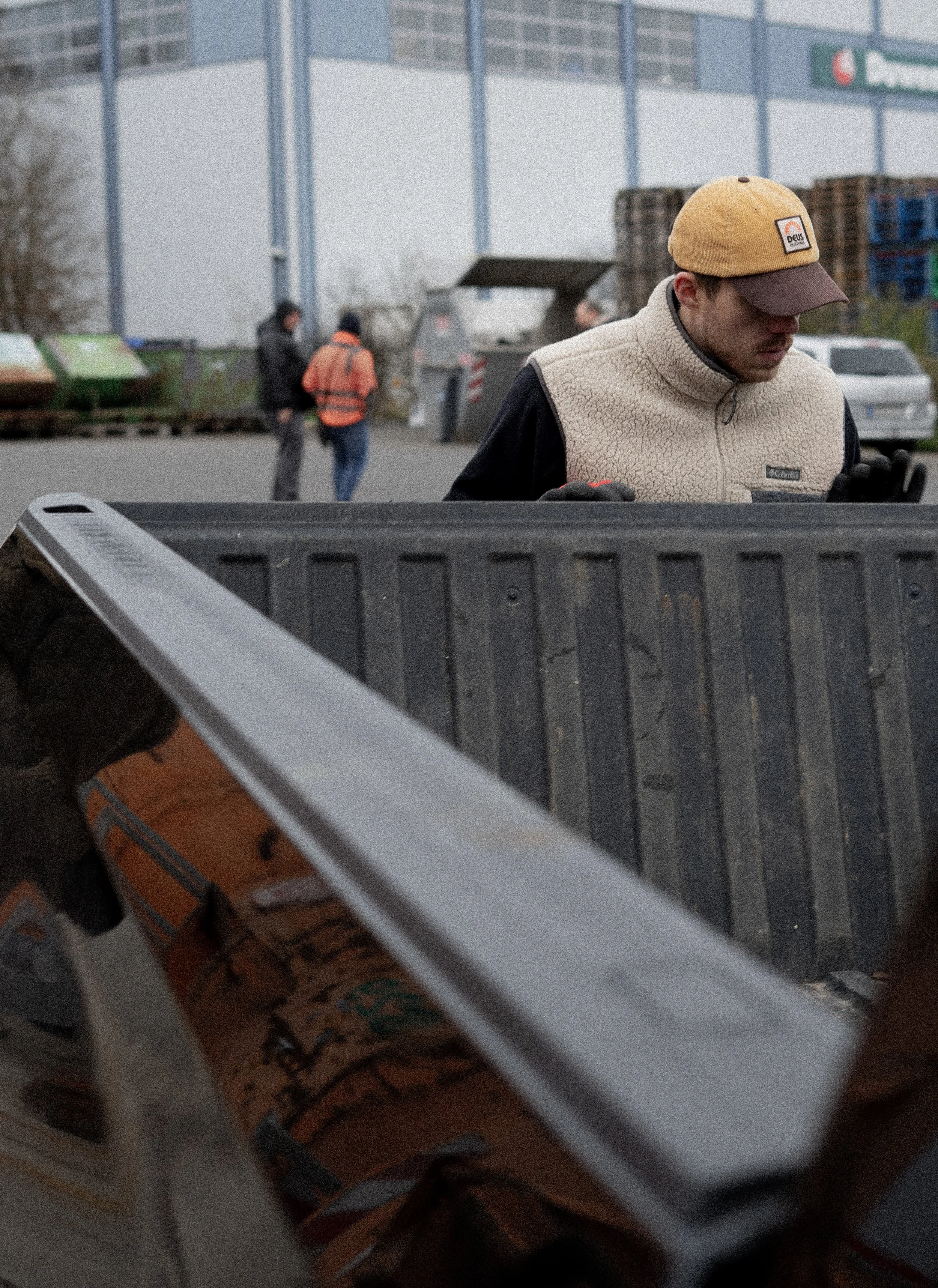 Junge Mann in beige Weste und Kappe steht an einem offenen Autokoffer im Freien, im Hintergrund mehrere Menschen und Industriehallen.
