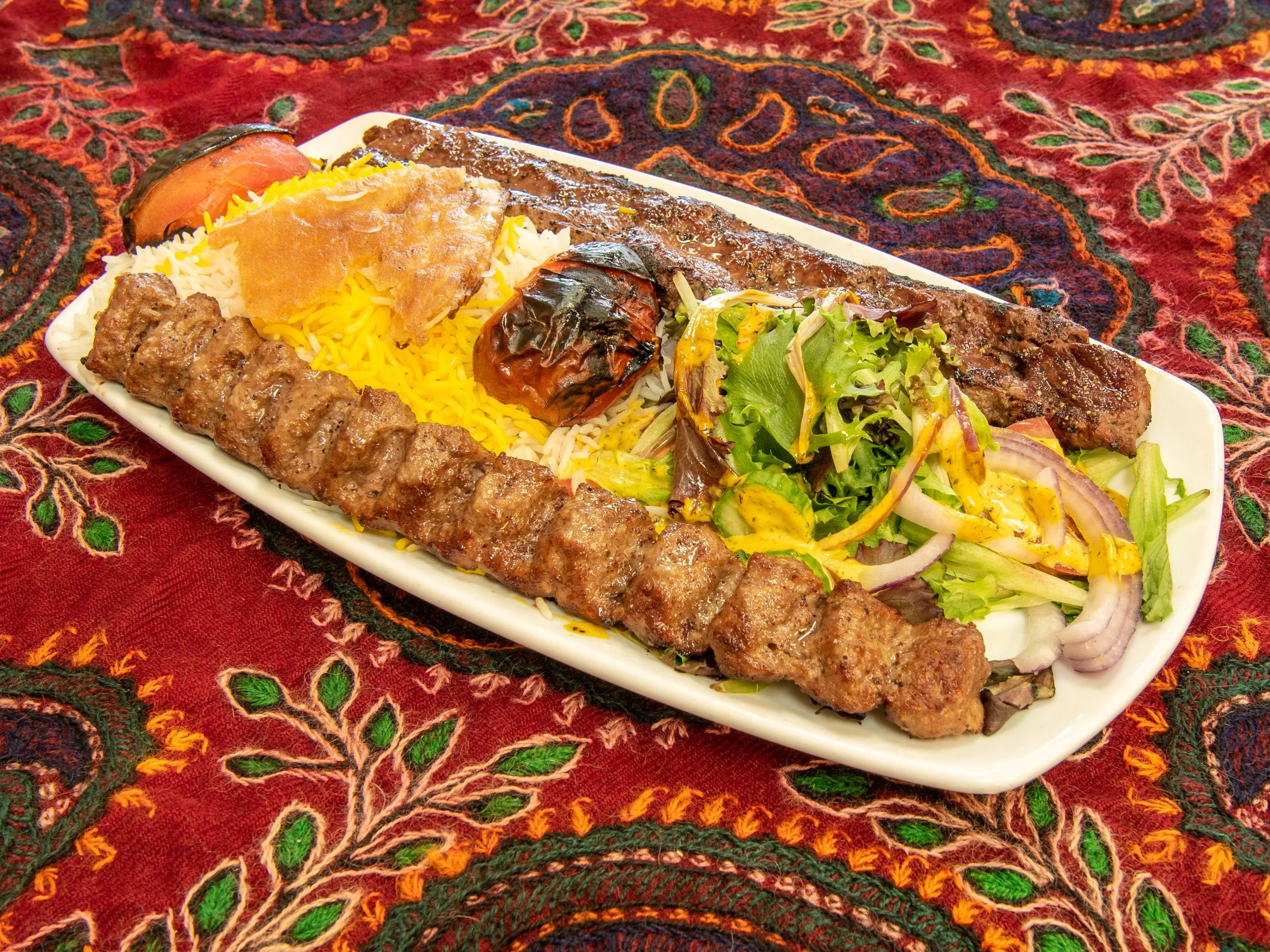 A plate of Middle Eastern food with grilled meats, rice, vegetables, and bread on a colorful fabric tablecloth.