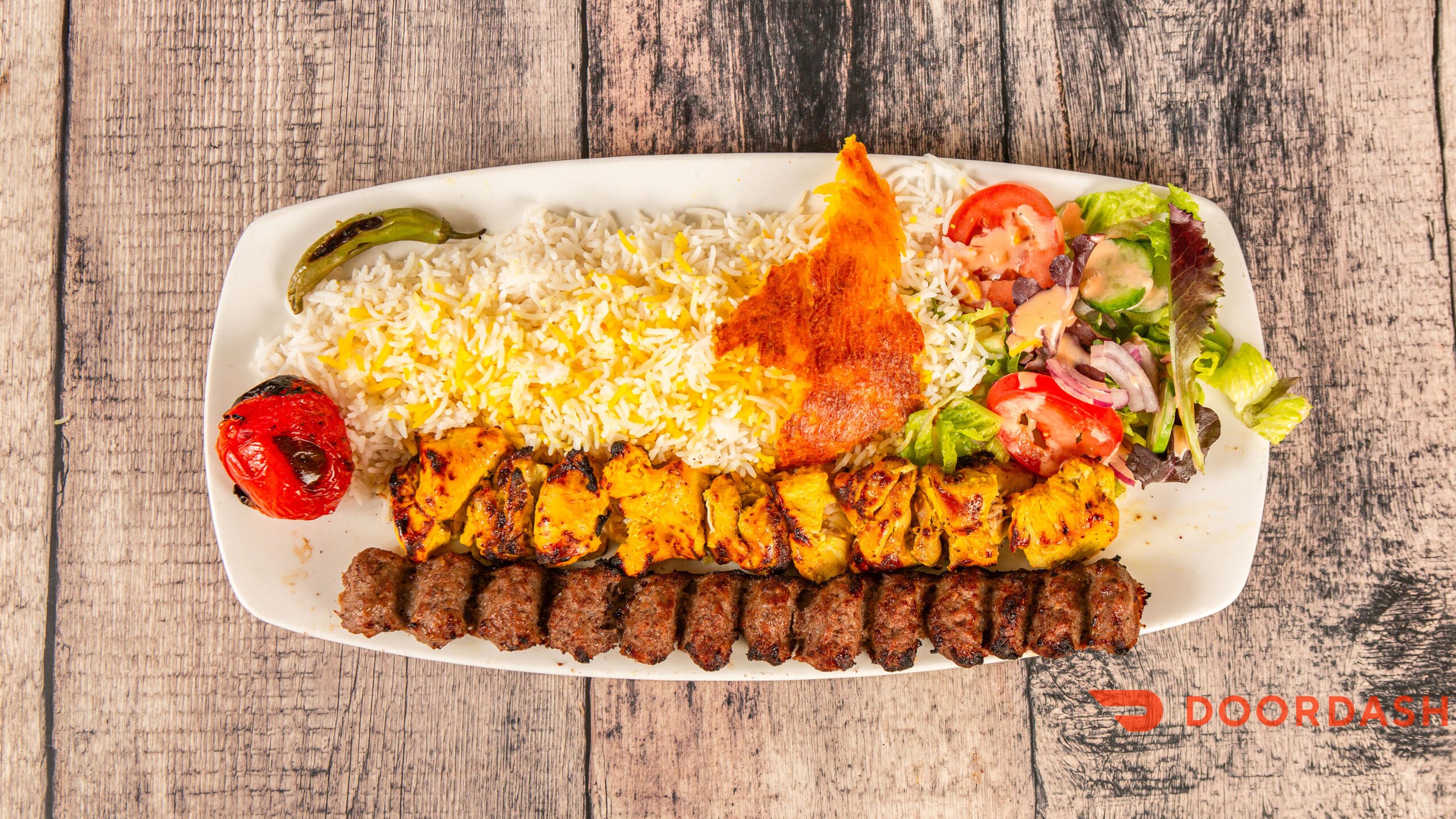 Plate of mixed grilled food including rice, grilled chicken, grilled beef kebab, grilled fish, a roasted tomato, and a salad with cherry tomatoes, cucumbers, and lettuce, on a wooden table.