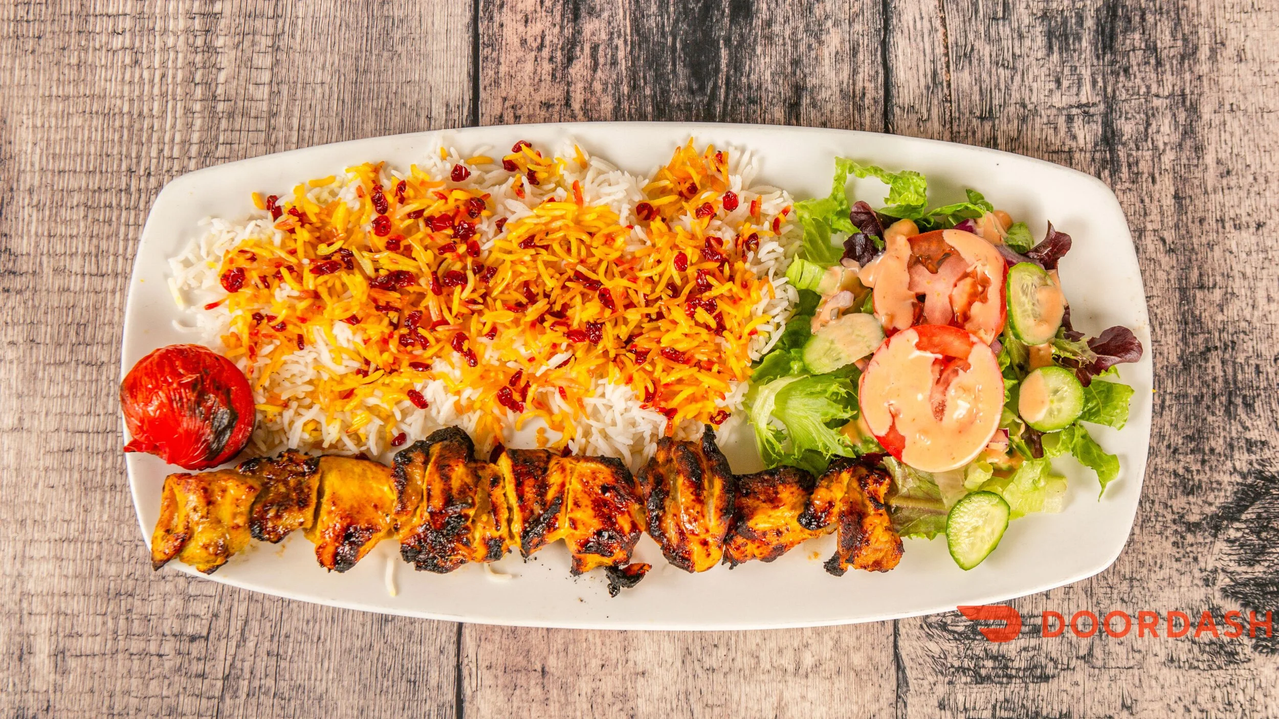 Plate of rice with saffron and tomato garnish, grilled chicken kebab, side salad with cucumbers, tomatoes, and dressing, and roasted tomato, on a wooden table.