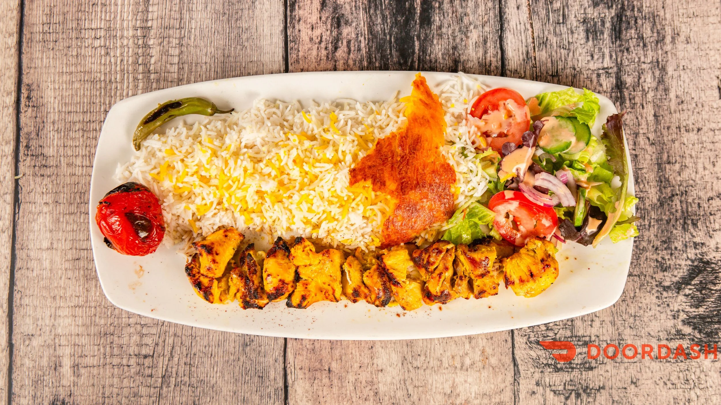 Plate of rice with yellow saffron threads, grilled chicken pieces, a piece of fried fish, grilled tomatoes, and a side salad with lettuce, cucumber, tomato, and onion with dressing, on a rustic wooden table.