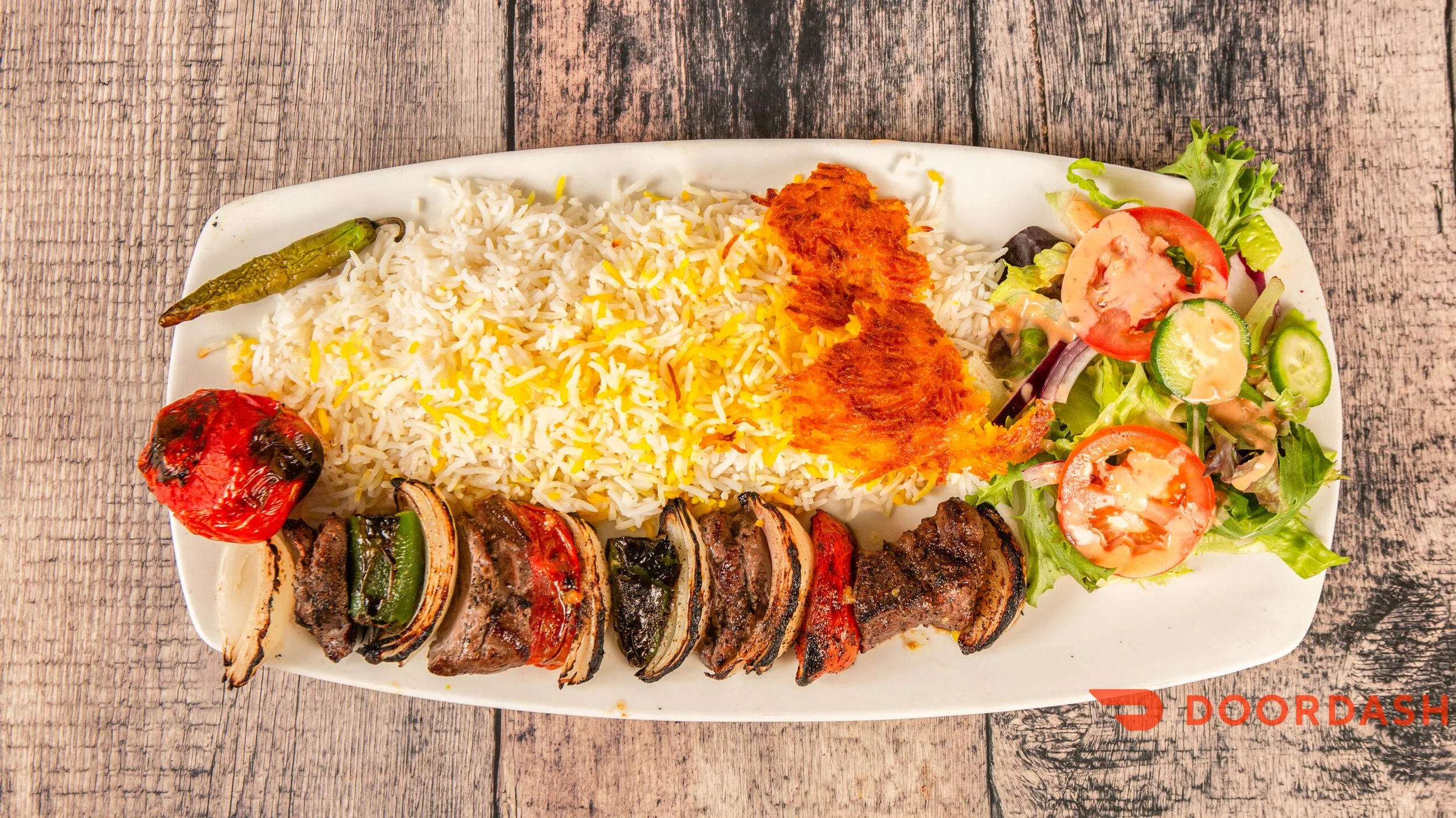 A plate of Persian rice with saffron, grilled meat and vegetables, a side salad with tomatoes, cucumbers, and lettuce, and a roasted tomato and green chili pepper on a wooden table.