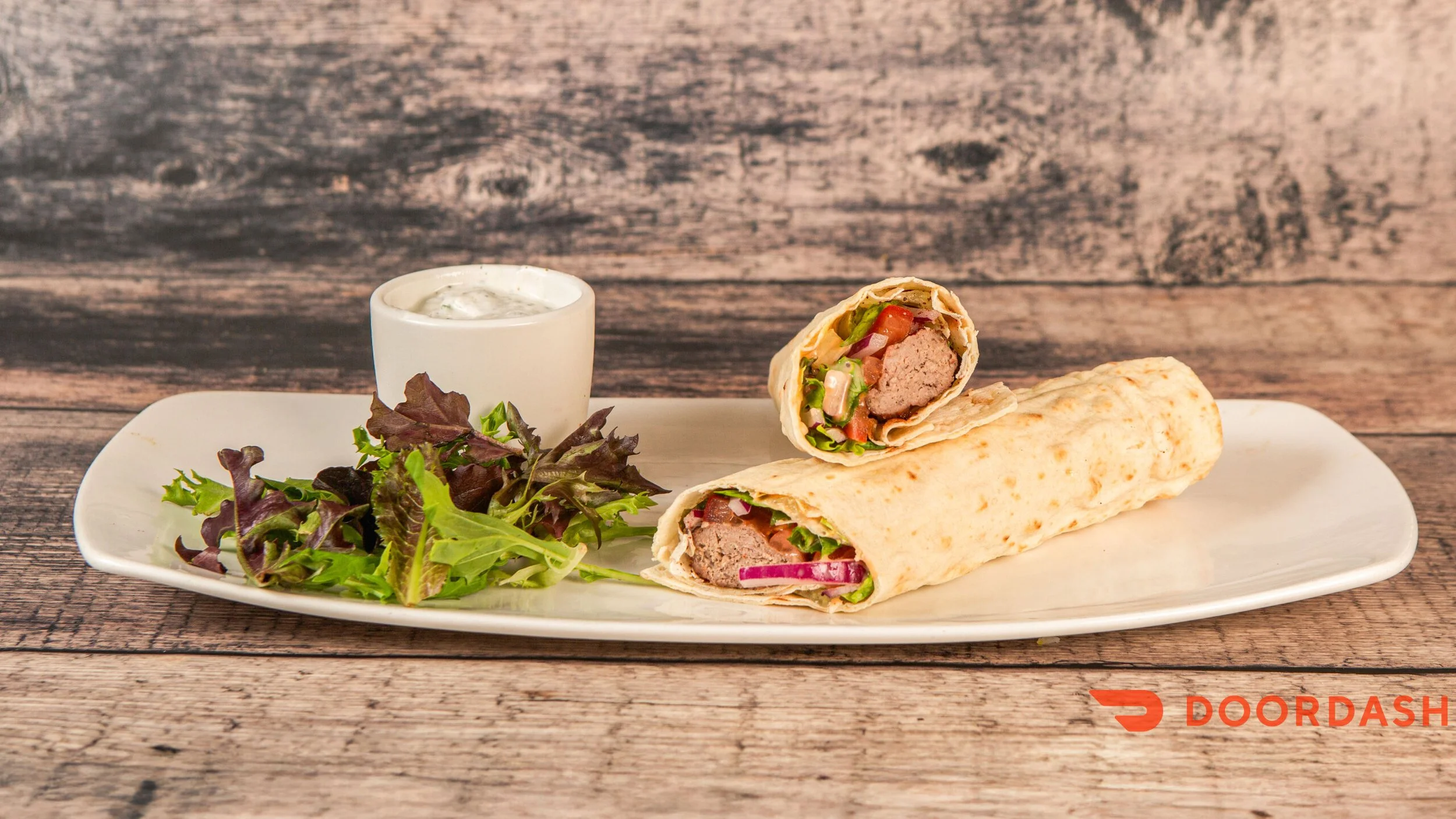 Plate with a wrap sandwich cut in half, a side salad, and a small cup of dressing on a wooden table.