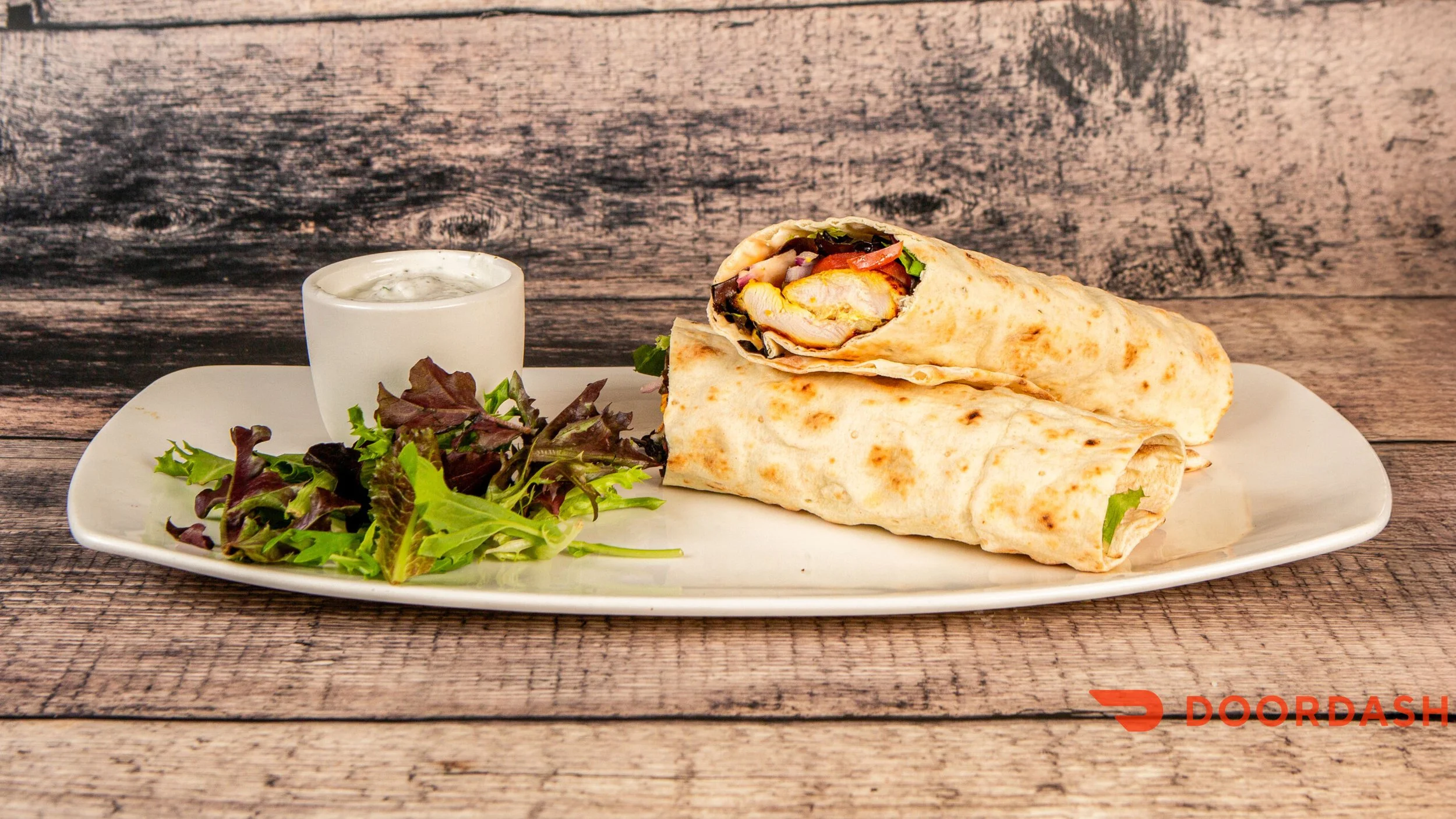 Two chicken wrap sandwiches on a white rectangular plate with a side of mixed greens and a small cup of creamy white dipping sauce, placed on a wooden surface with a rustic background.