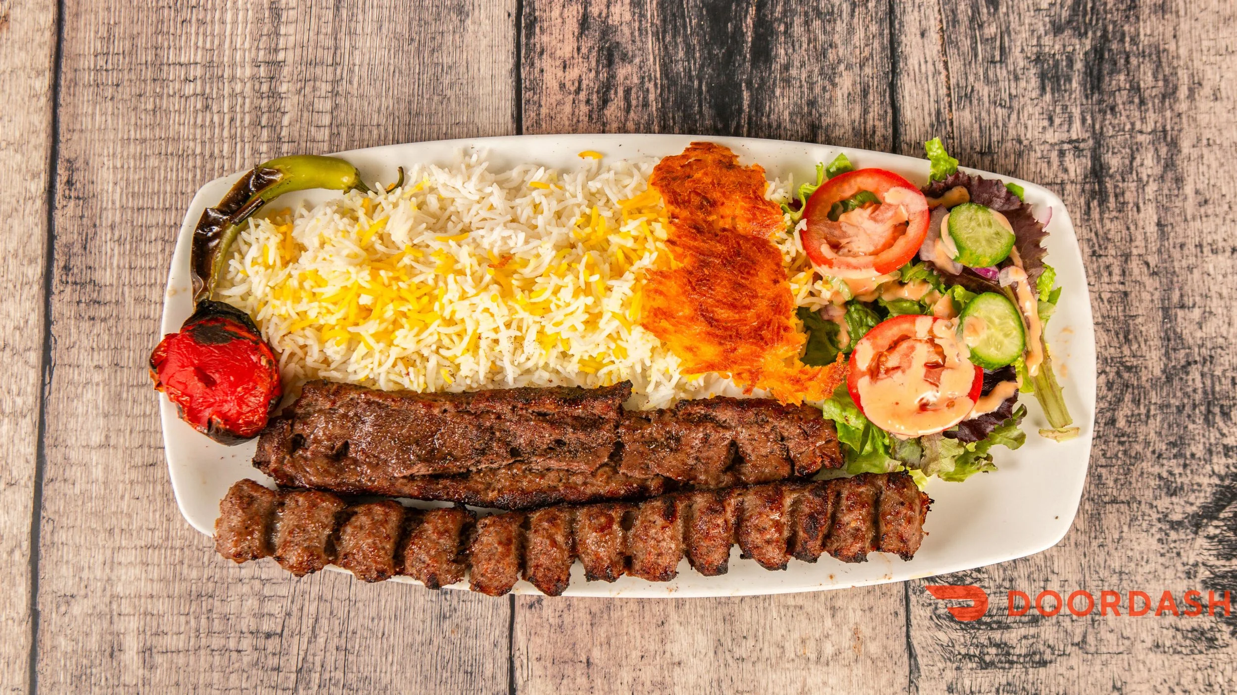 Plate of rice with yellow saffron threads, grilled kebabs, grilled vegetables, and a side salad with tomatoes, cucumbers, and lettuce, on a wooden table.