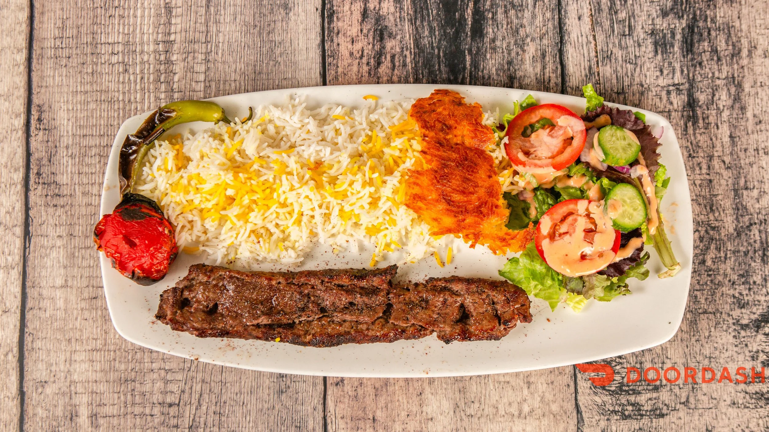 Plate of rice with yellow and white grains, a grilled kebab, two pieces of fried plantain, grilled tomato, two pieces of grilled green chili, and a mixed salad with tomato, cucumber, lettuce, and dressing on a wooden table.