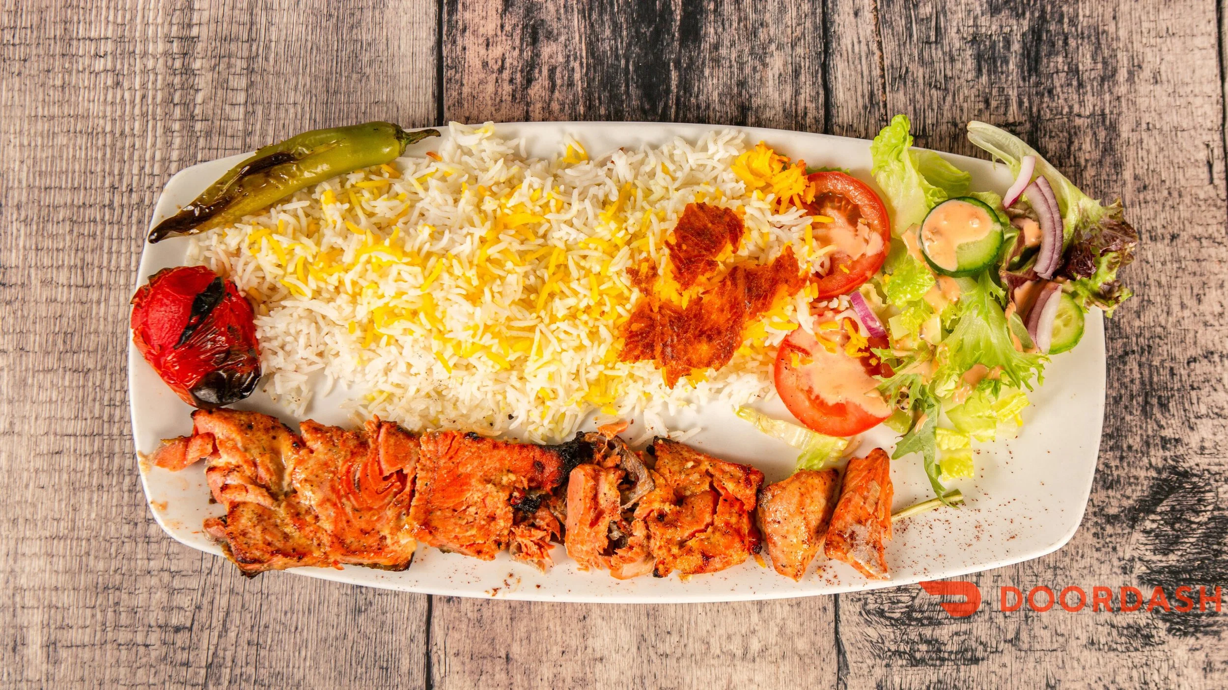 Plate of rice with yellow saffron, grilled tomato, green chili, grilled chicken pieces, and side salad with cucumber, tomato, lettuce, onion, and dressing. Volcanic rock on the side, on a wooden table.