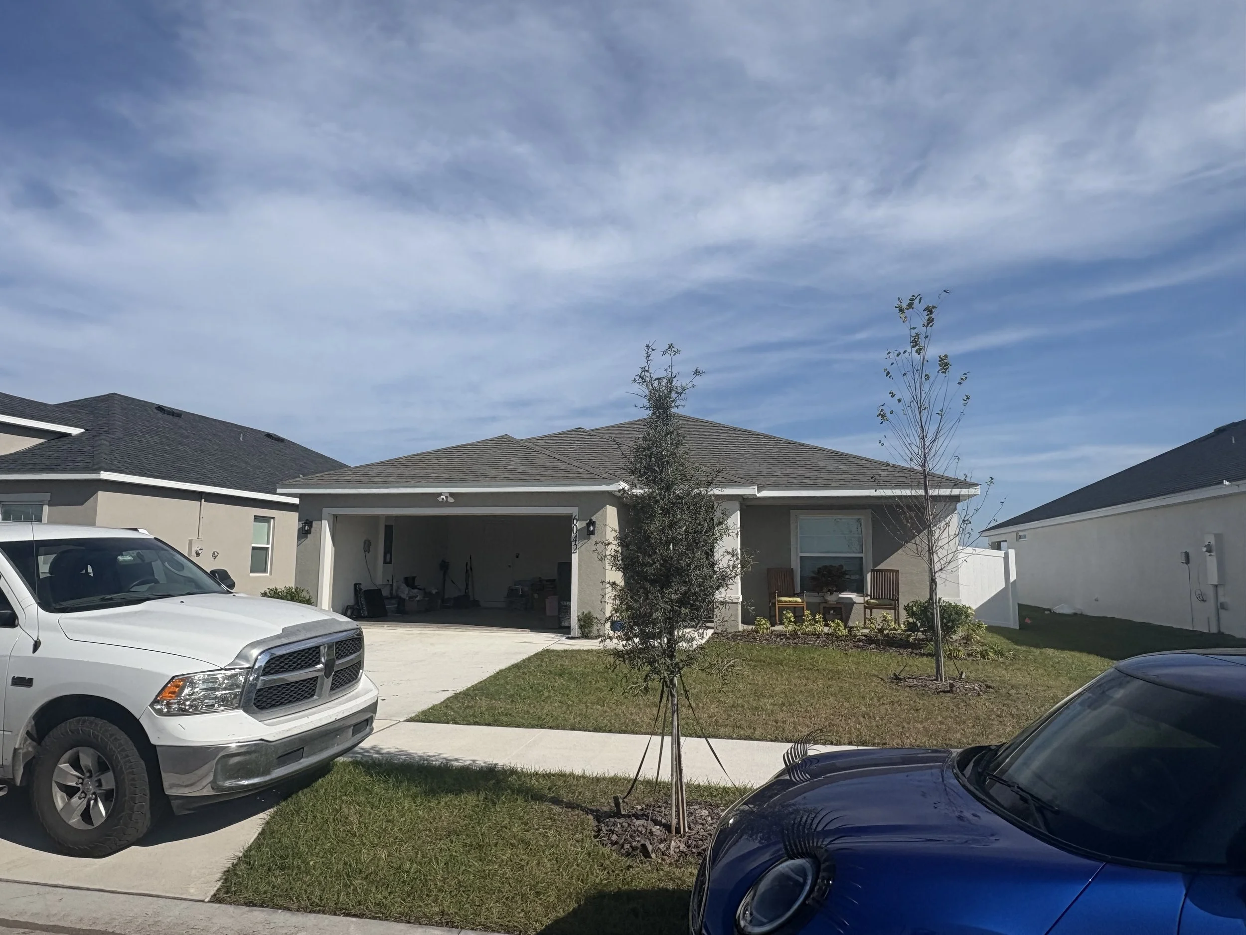 Front yard of a suburban house with two parked cars, trees, and a porch with chairs under a cloudy sky.