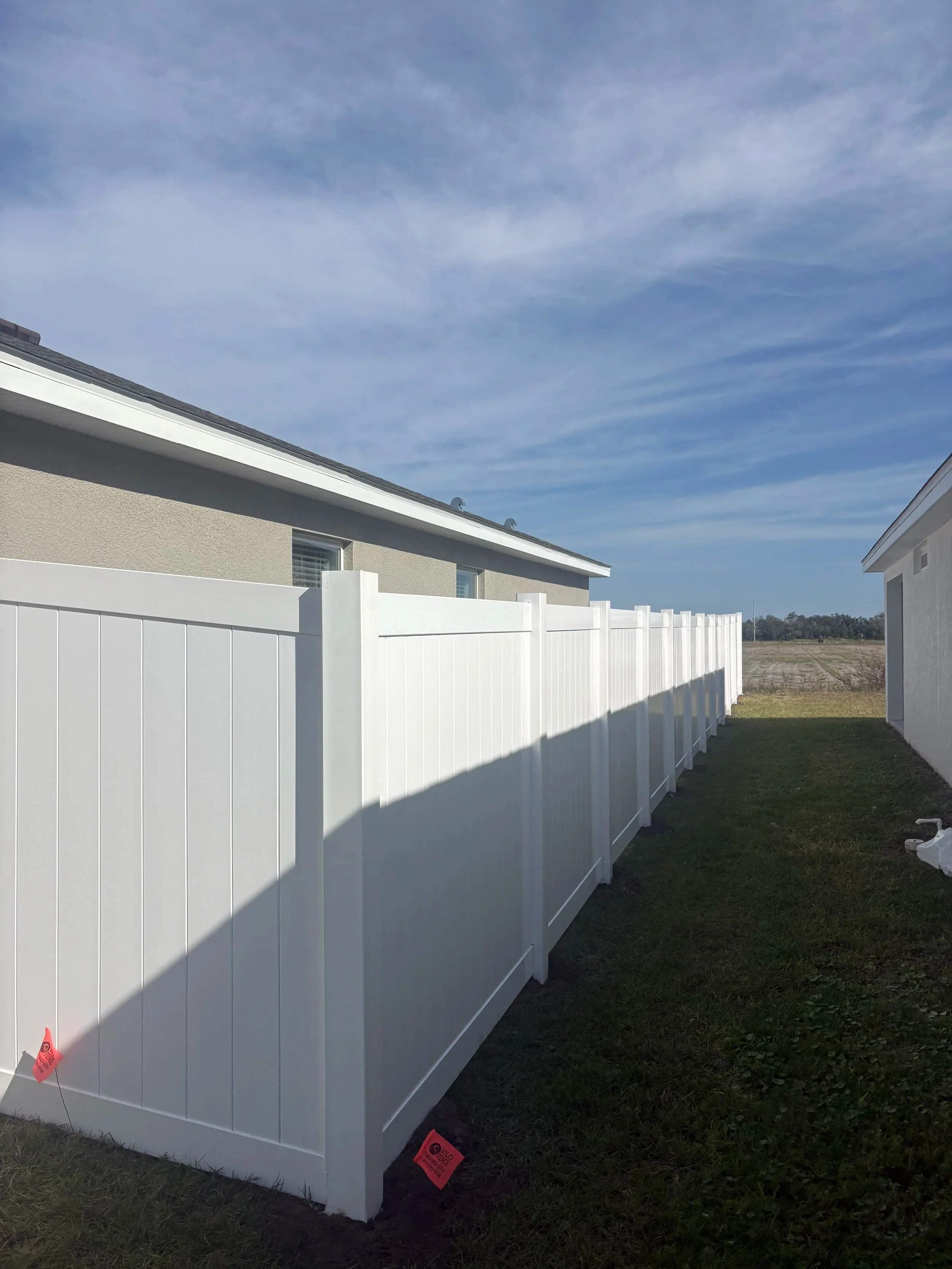 White privacy fence running between two houses on a grassy lot, with a partly cloudy blue sky overhead.