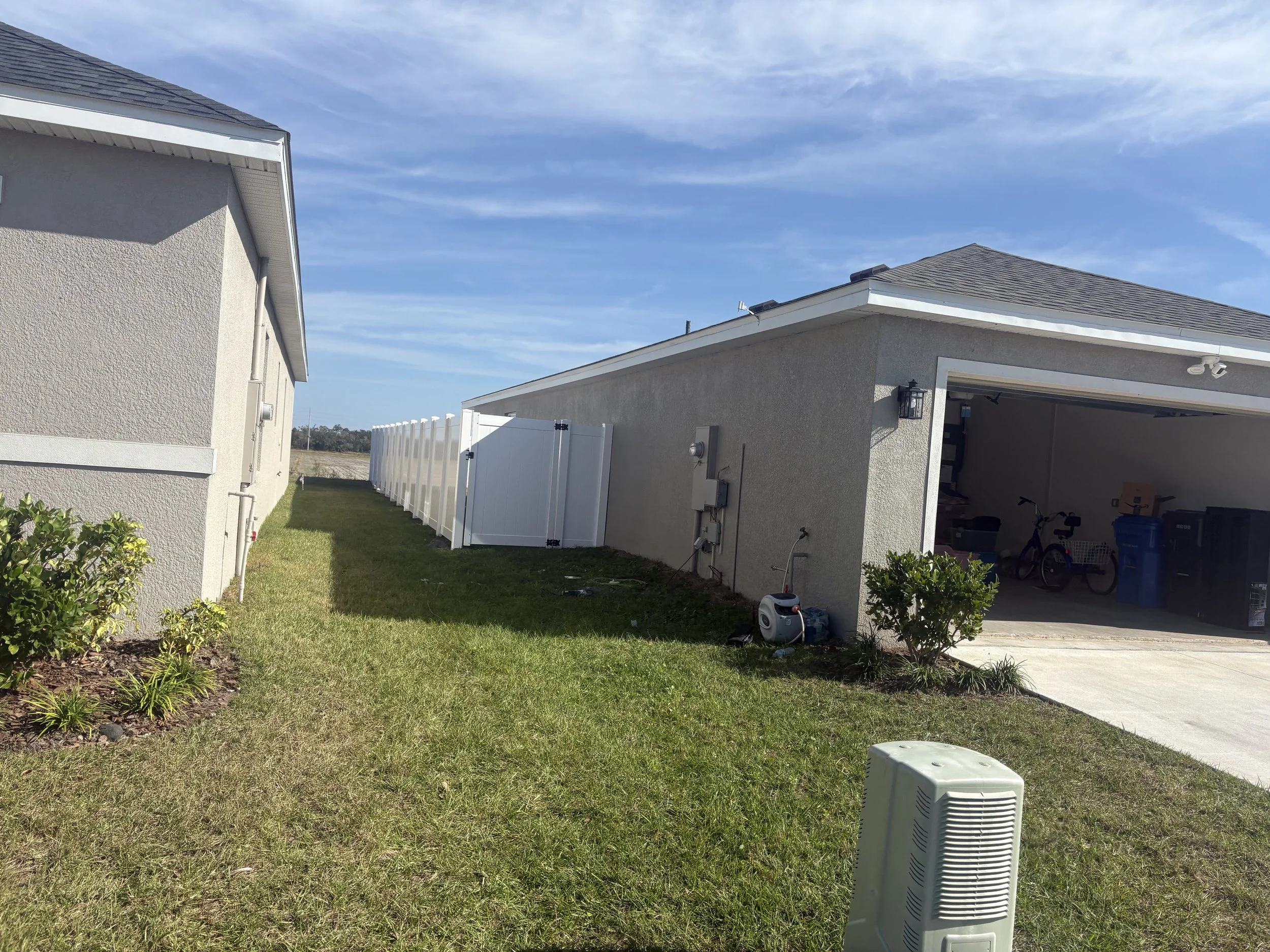 Side view of a single-story house with a garage, a white fence, green grass, and a clear blue sky.