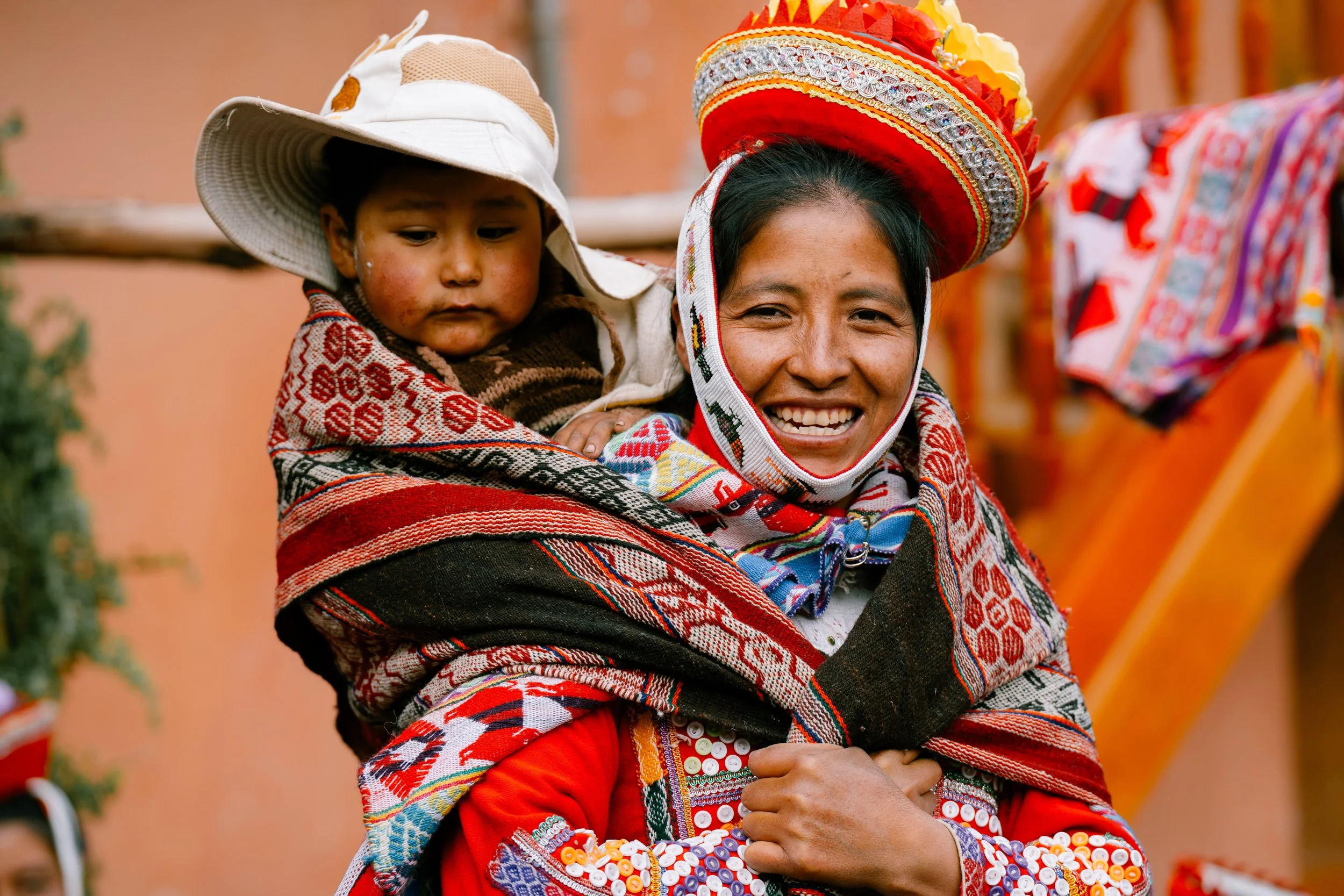 A smiling woman wearing traditional colorful clothing and a large decorated hat, carrying a baby on her back wrapped in a patterned fabric, outdoors with a blurred background.