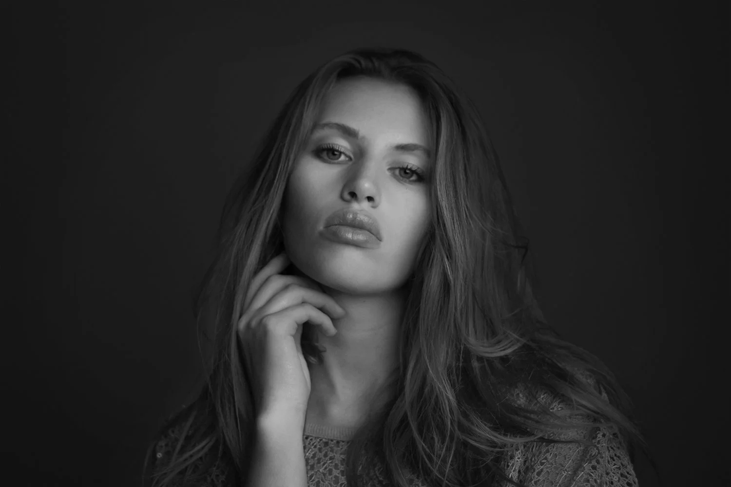 Black and white portrait of a young woman with long, wavy hair, lightly touching her chin, against a dark background.