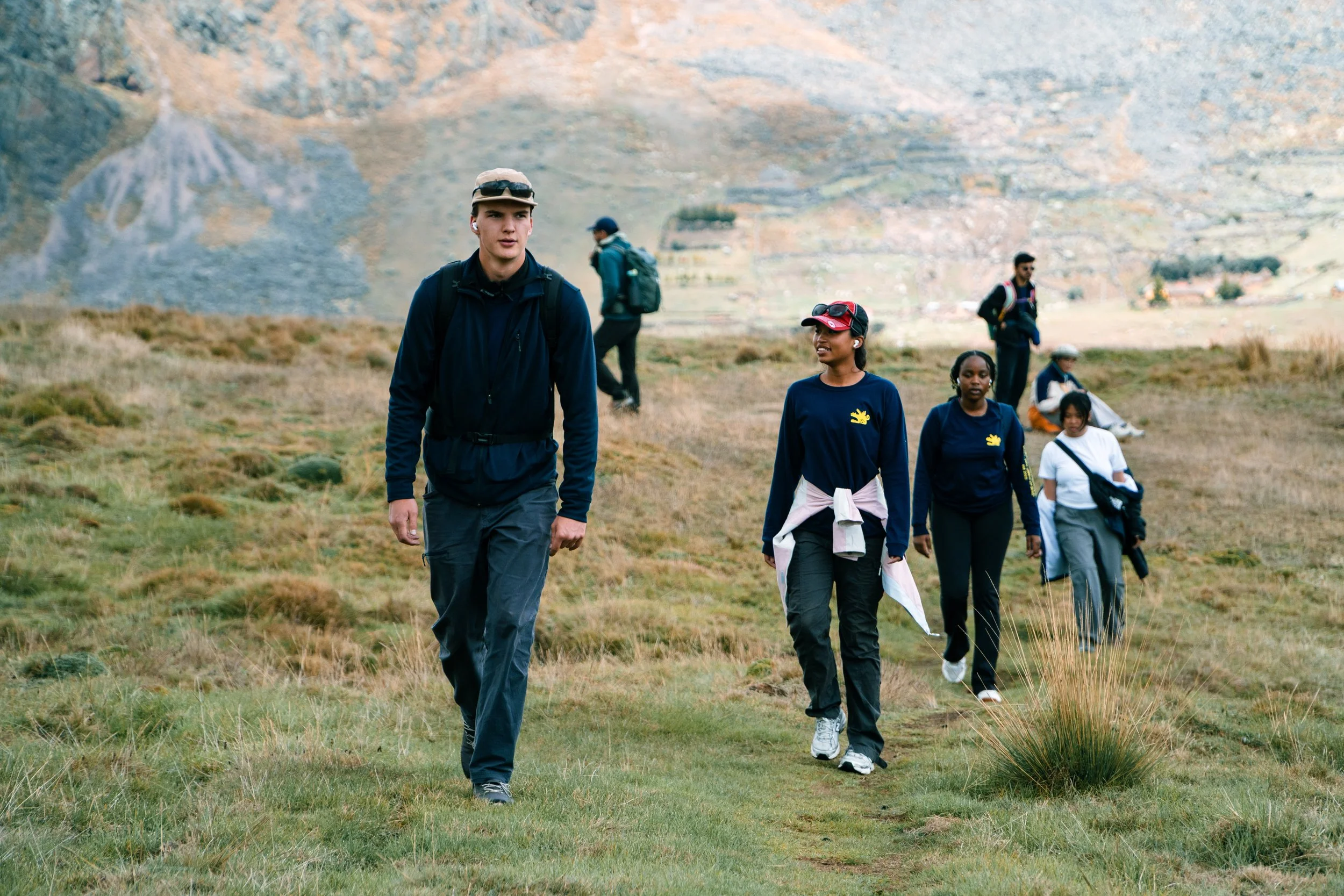 A group of people hiking in a mountainous area, with grassy terrain and rocky cliffs in the background.