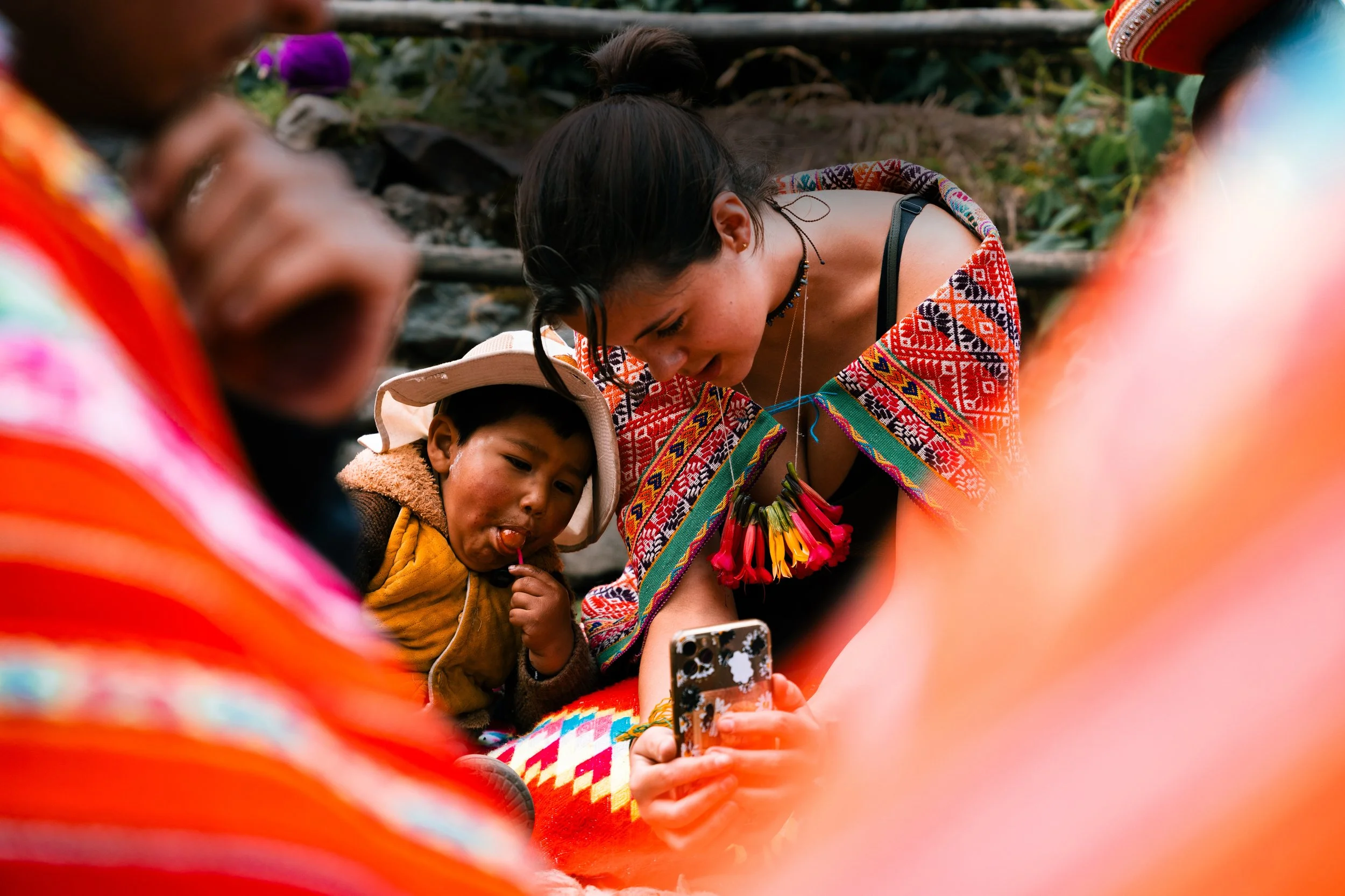 A woman with long dark hair and colorful traditional clothing taking a selfie on her phone while a young boy in a hat and yellow jacket looks at the camera, surrounded by blurred people.