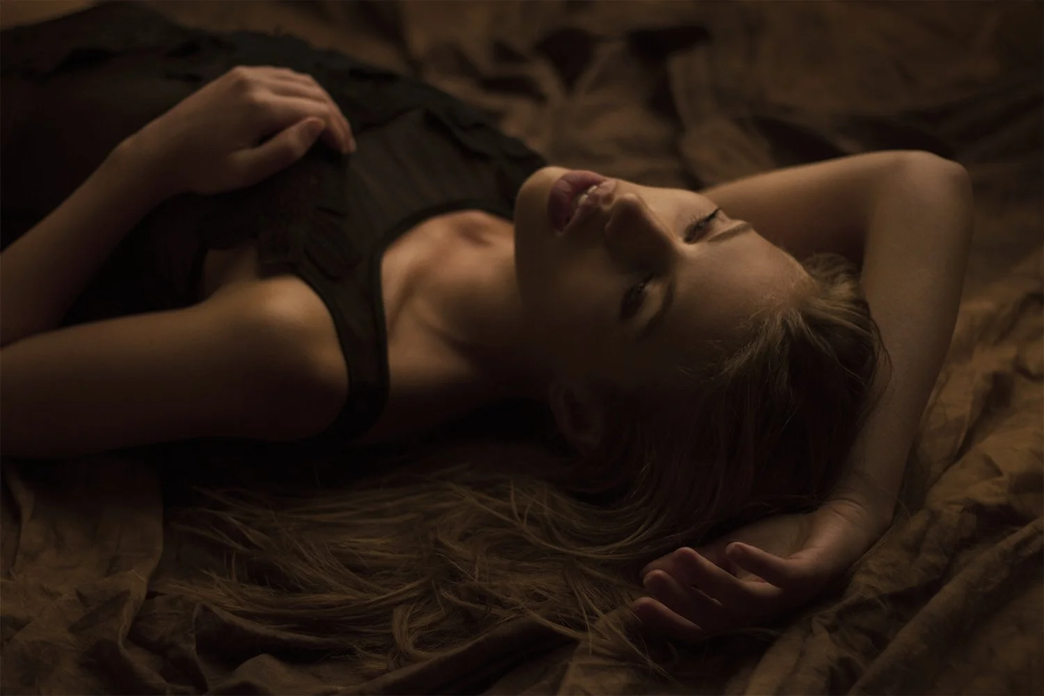 A woman with long hair lying on a bed with brown sheets, wearing a black sleeveless top, her eyes closed, and one hand resting on her stomach.