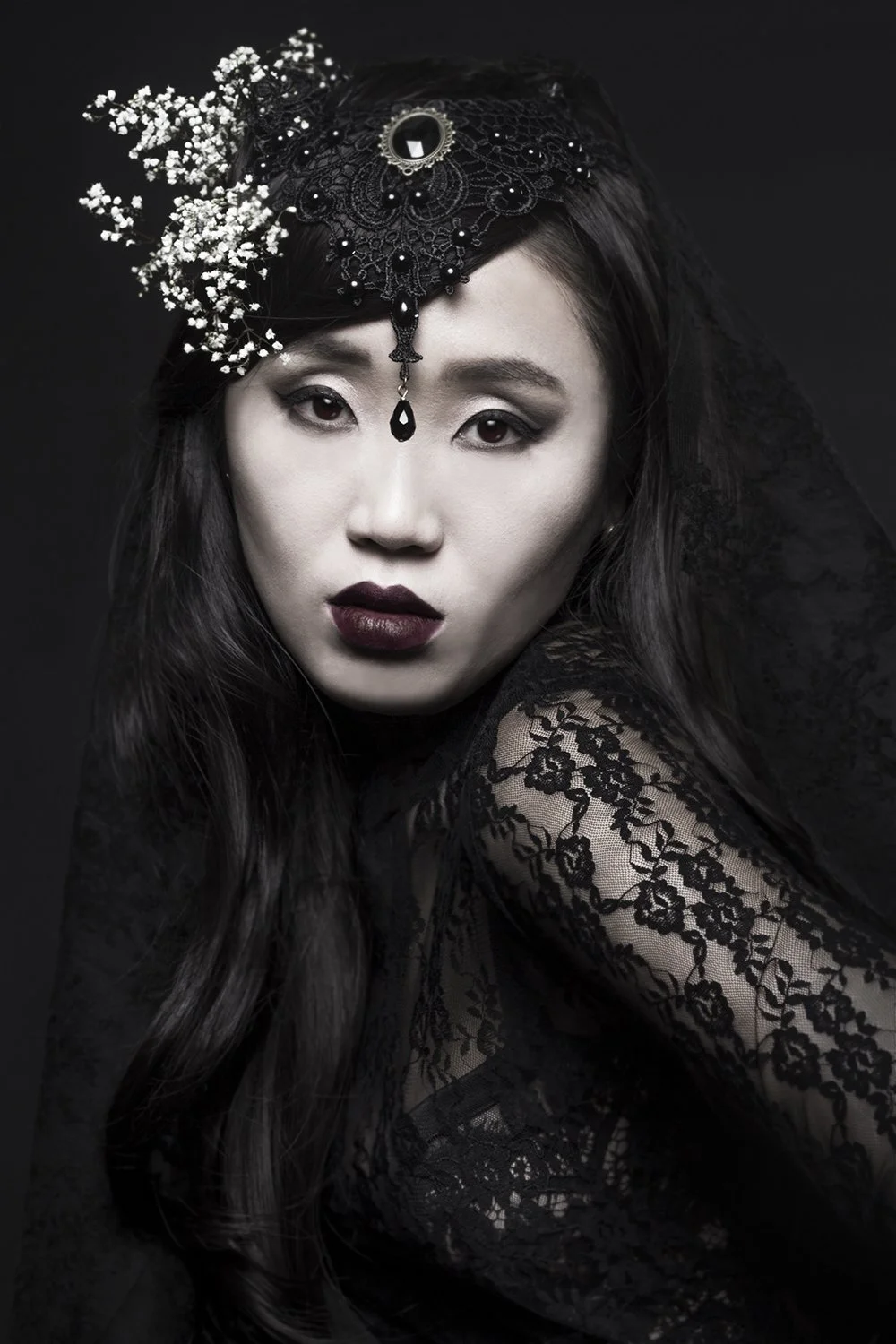 A woman with dark hair and makeup, wearing black lace clothing and ornate black lace headpiece with floral accents and a pendant, against a dark background.