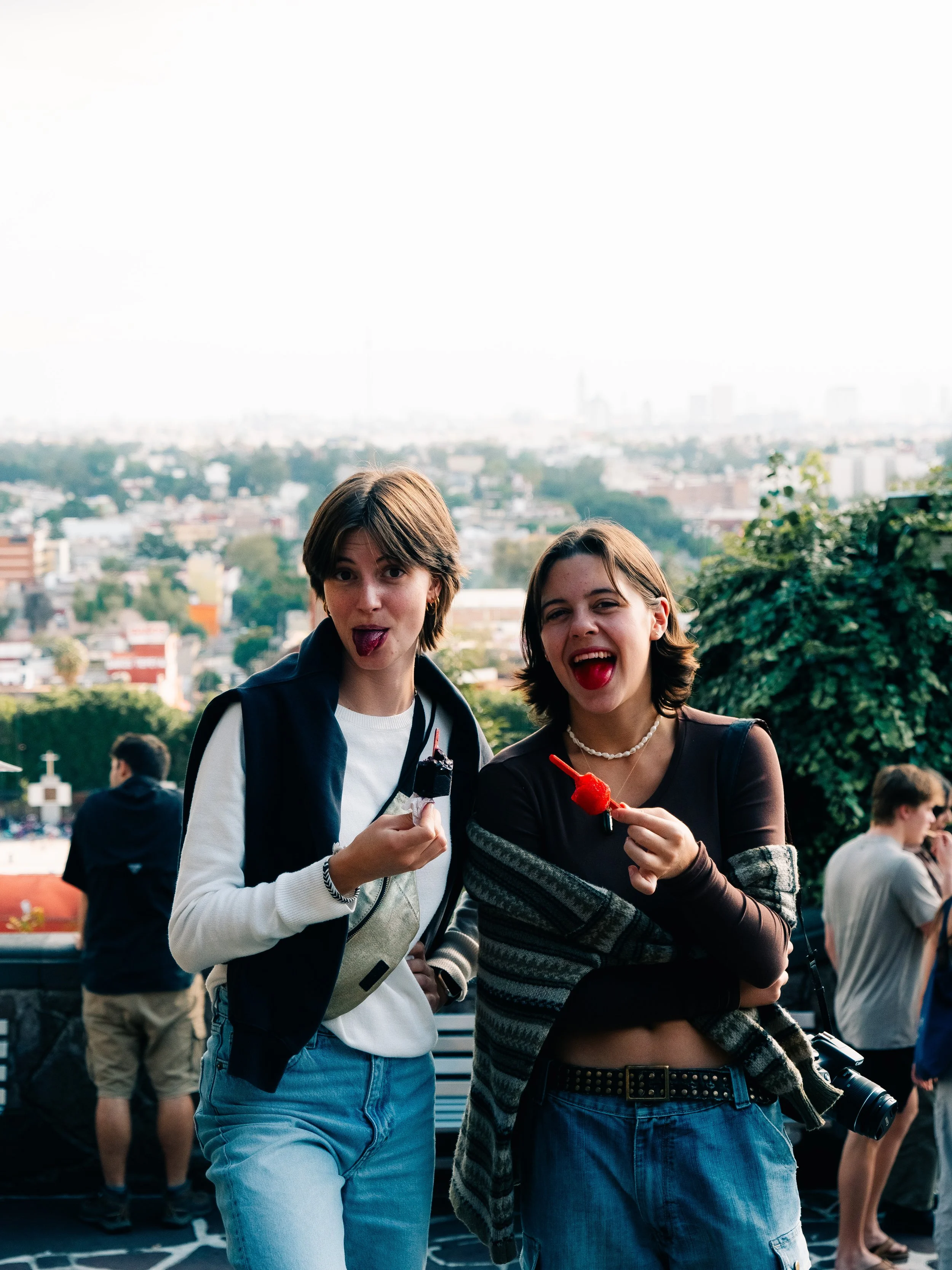 Two young women standing outdoors, smiling and holding colorful snacks, with a cityscape in the background.