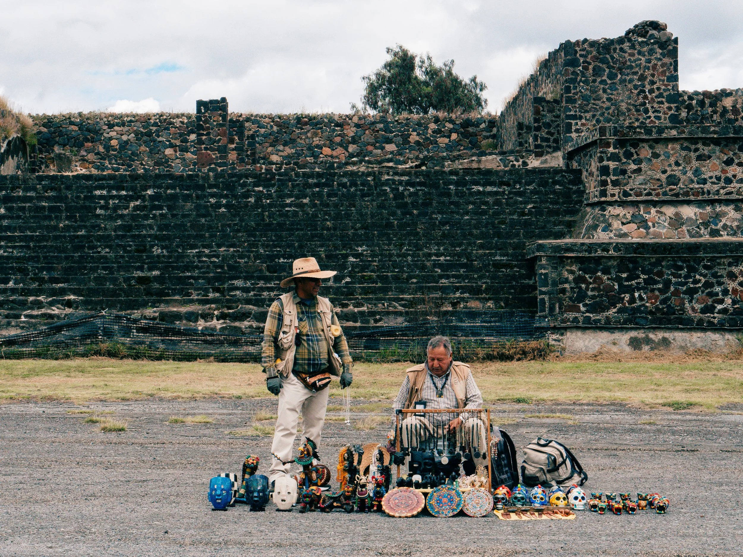 Two street vendors selling handmade colorful masks, jewelry, and souvenirs outdoors in front of an ancient stone archaeological site, with cloudy sky and trees in the background.