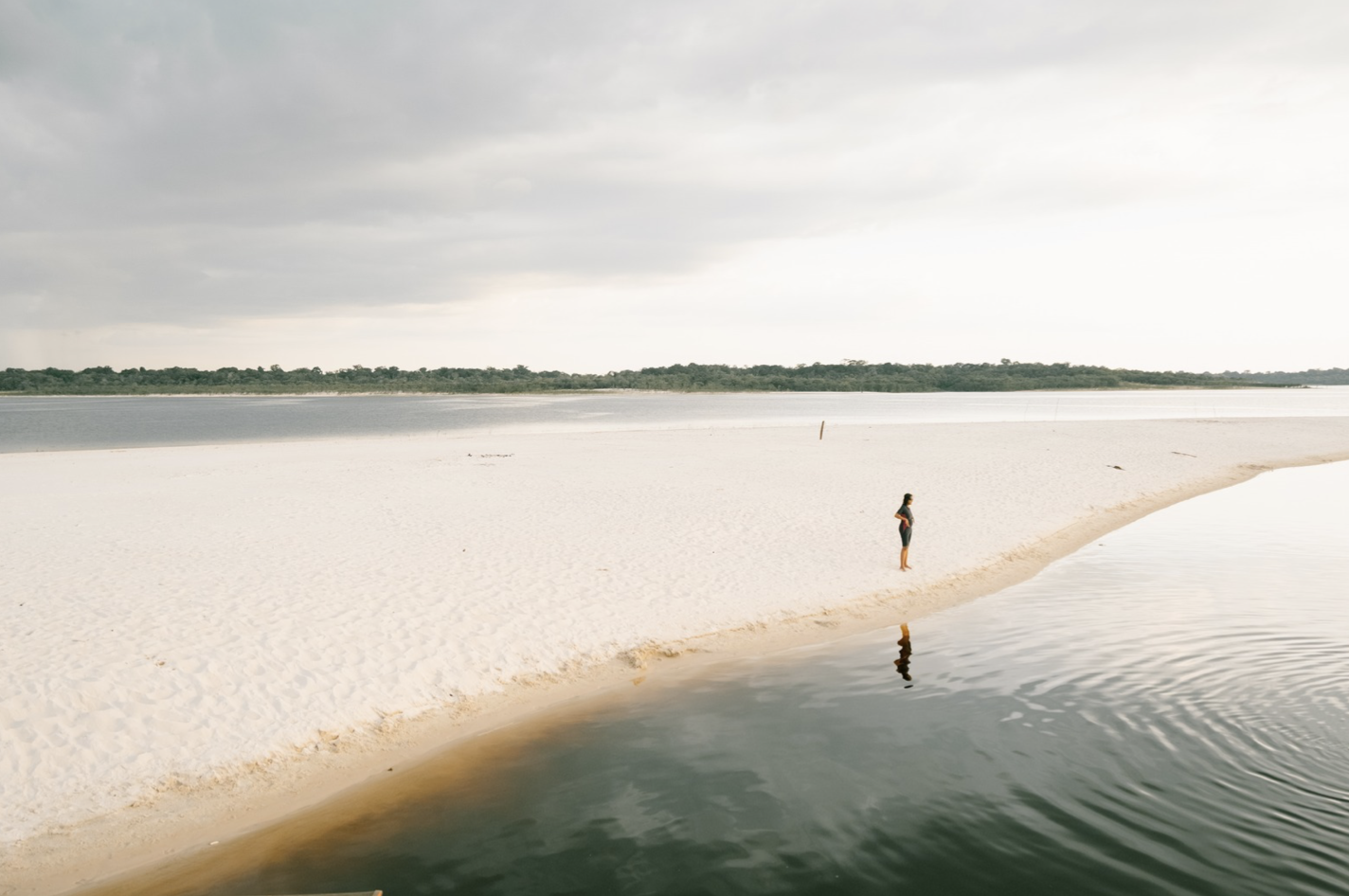 A person standing on a sandy beach near calm water, with a distant tree-lined horizon under cloudy sky.