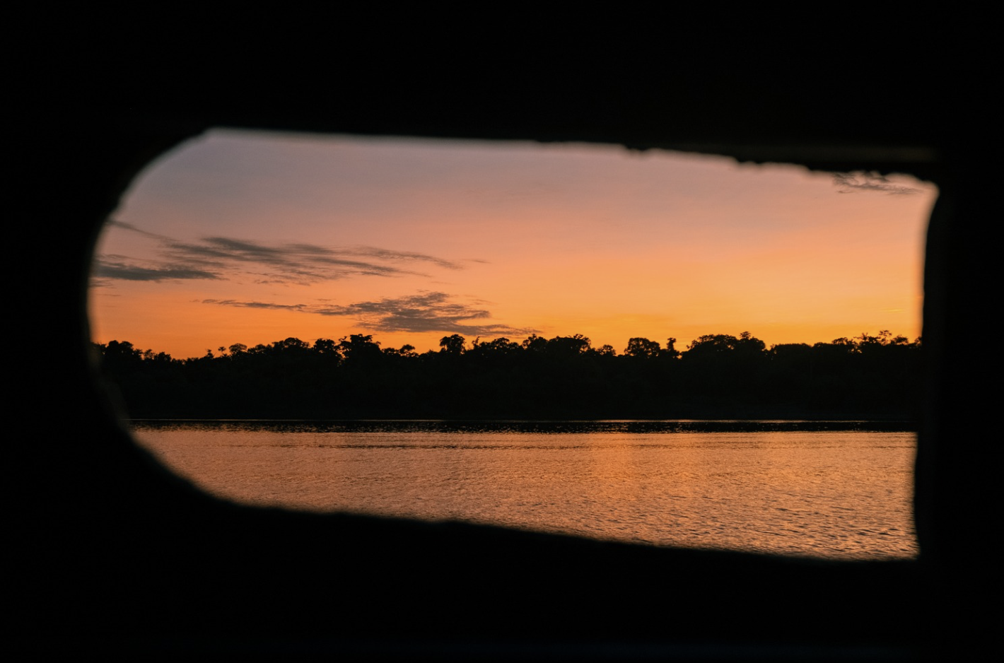 Sunset over a body of water, viewed through a rectangular opening with curved edges, with silhouetted trees on the horizon.