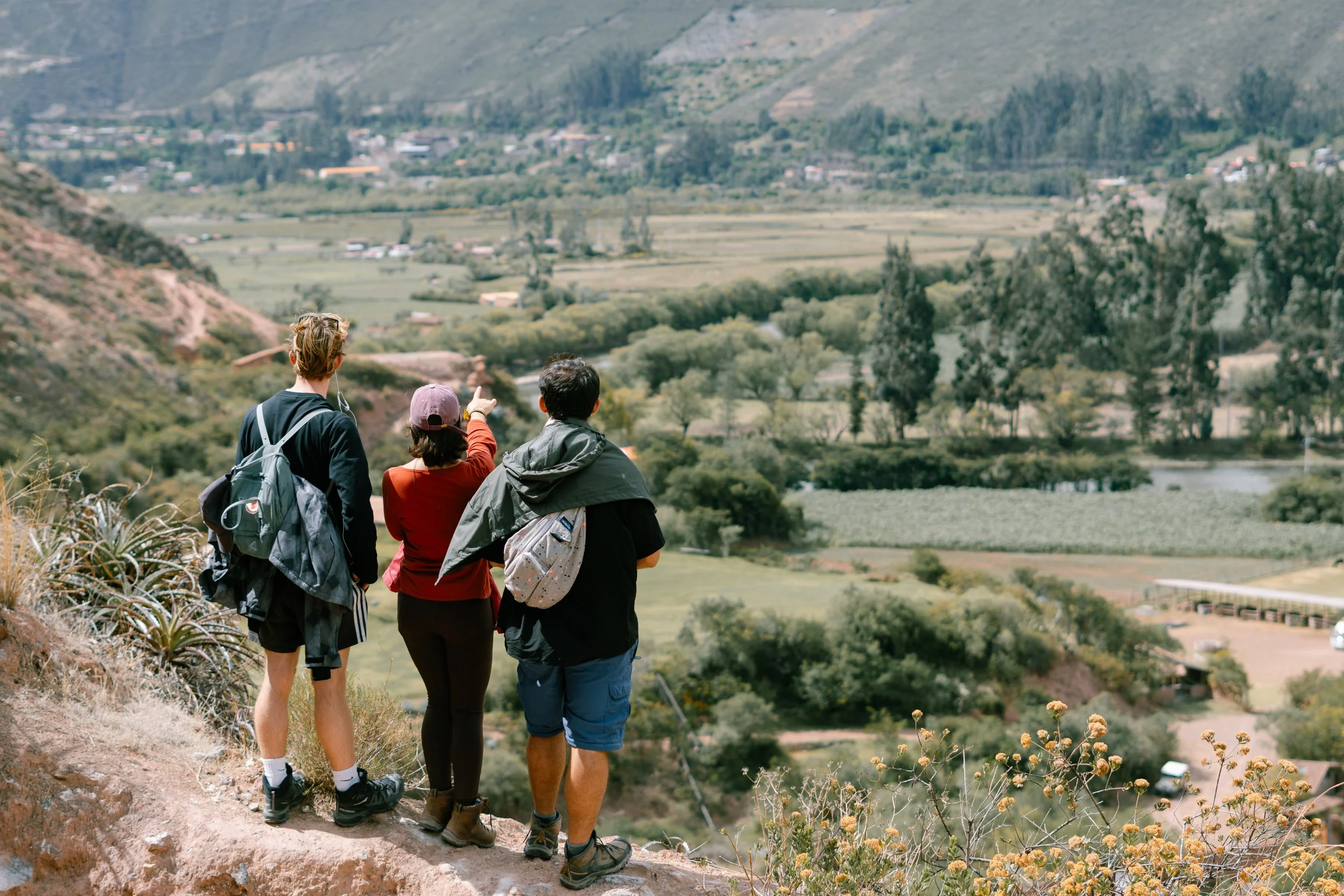 Three hikers on a trail overlooking a green valley with trees, fields, and hills in the distance.