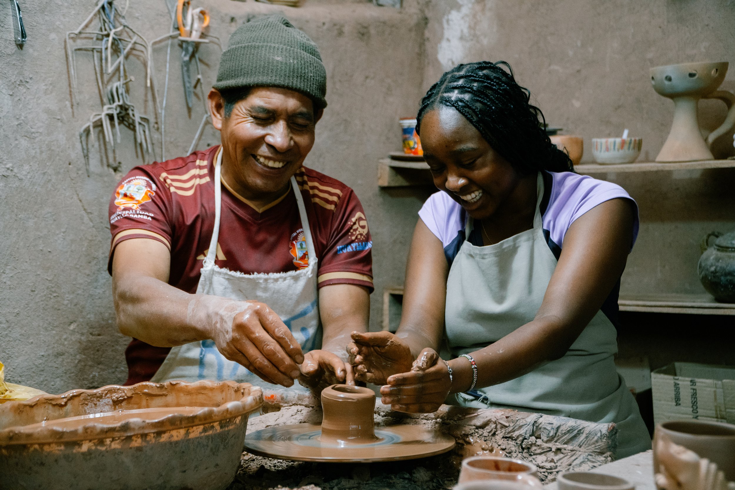 Two people, a man and a woman, are smiling and working together to shape clay on a pottery wheel in a rustic workshop. They appear happy and engaged in the activity.