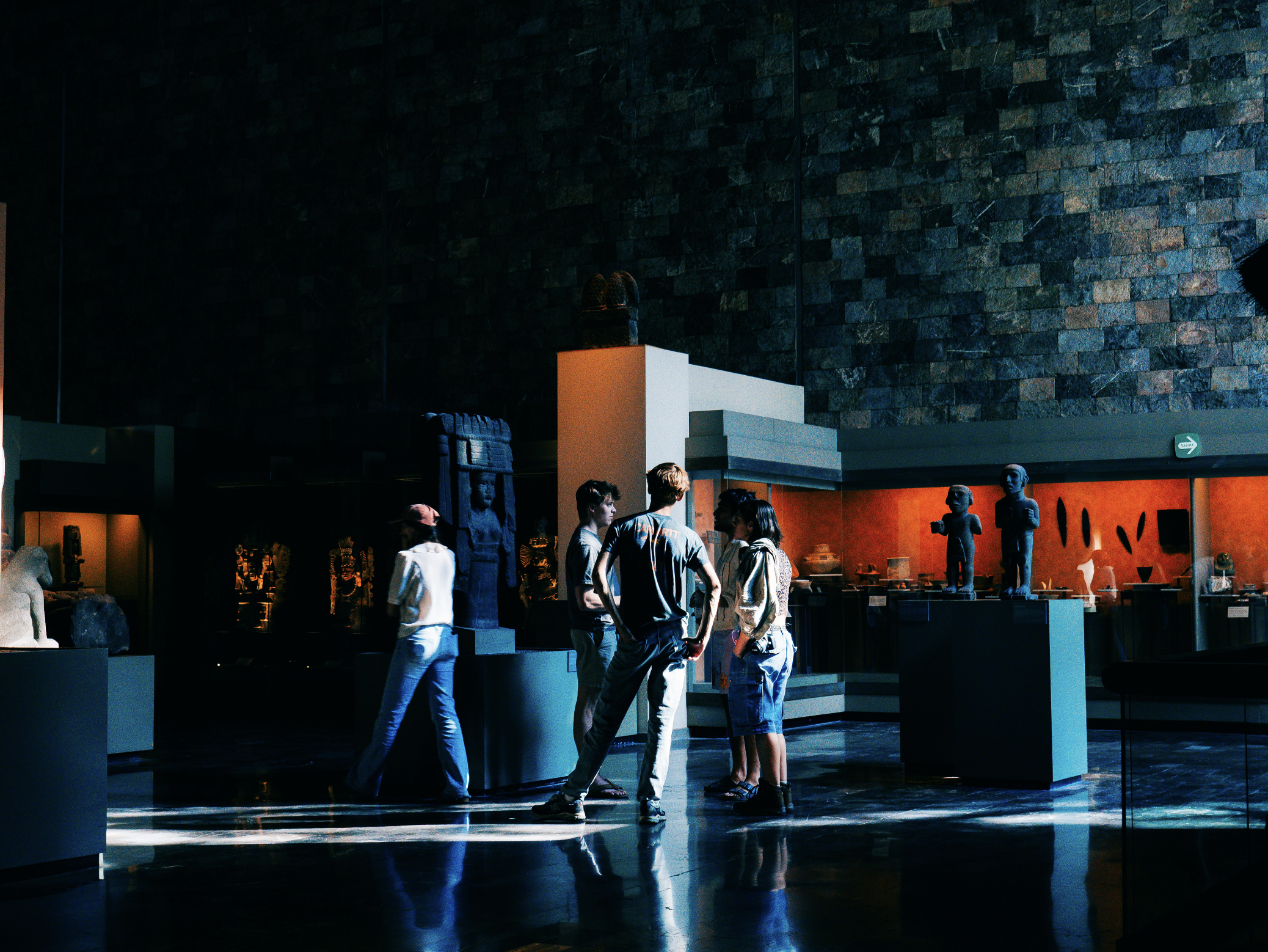 A group of young people standing and talking in a museum gallery with glass display cases and ancient artifacts, including sculptures and statues, illuminated by soft lighting.