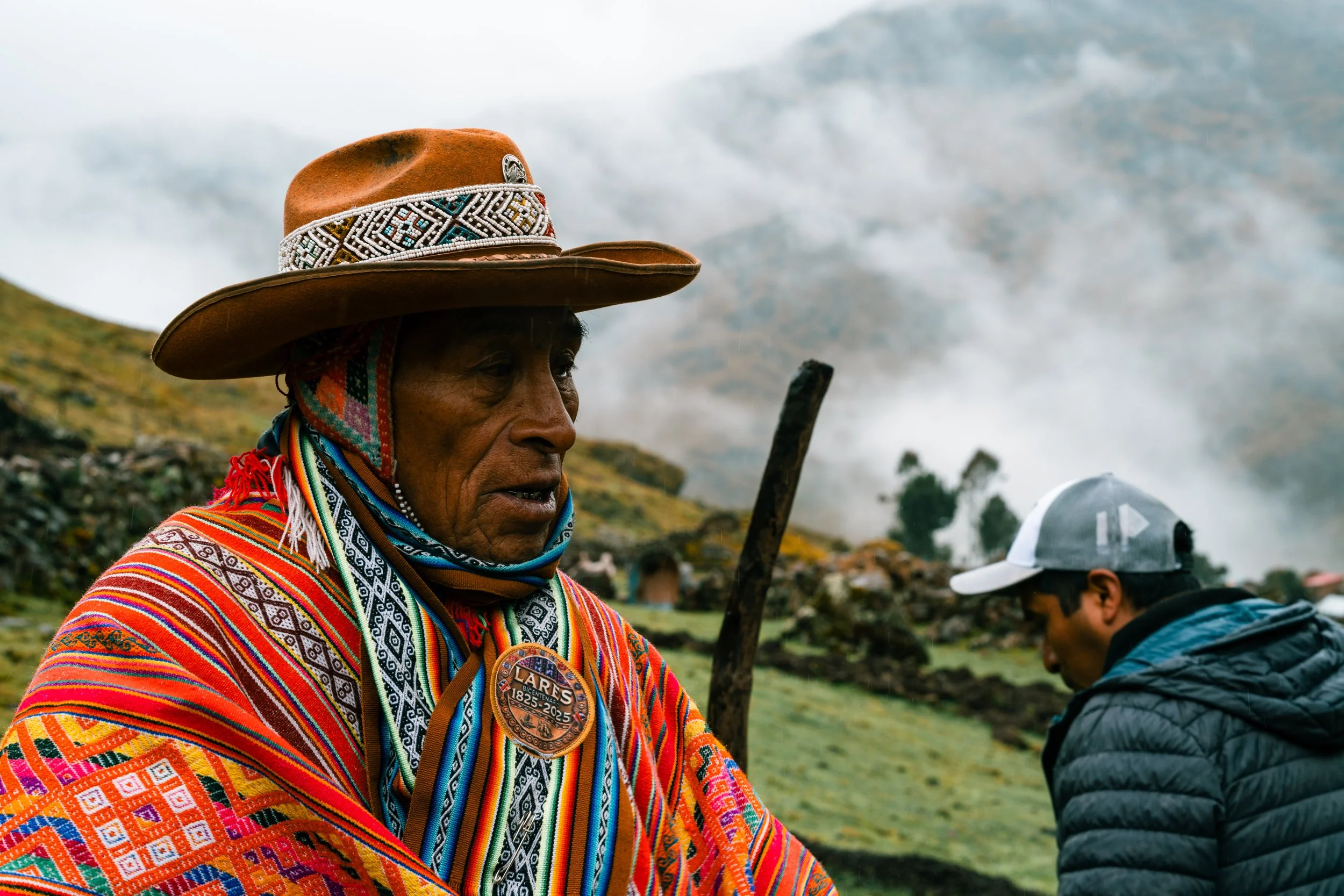 An indigenous woman wearing traditional colorful woven clothing, a large hat with decorative band, and a medallion, standing outdoors on a hillside with cloudy sky and a man in a cap and jacket in the background.