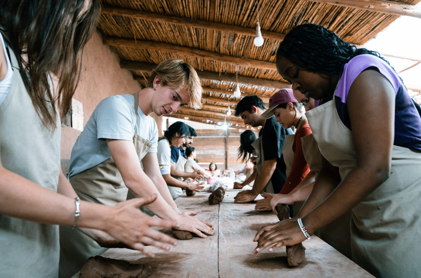 Group of diverse people working together on a pottery project at a long wooden table in a rustic workshop with a straw ceiling.