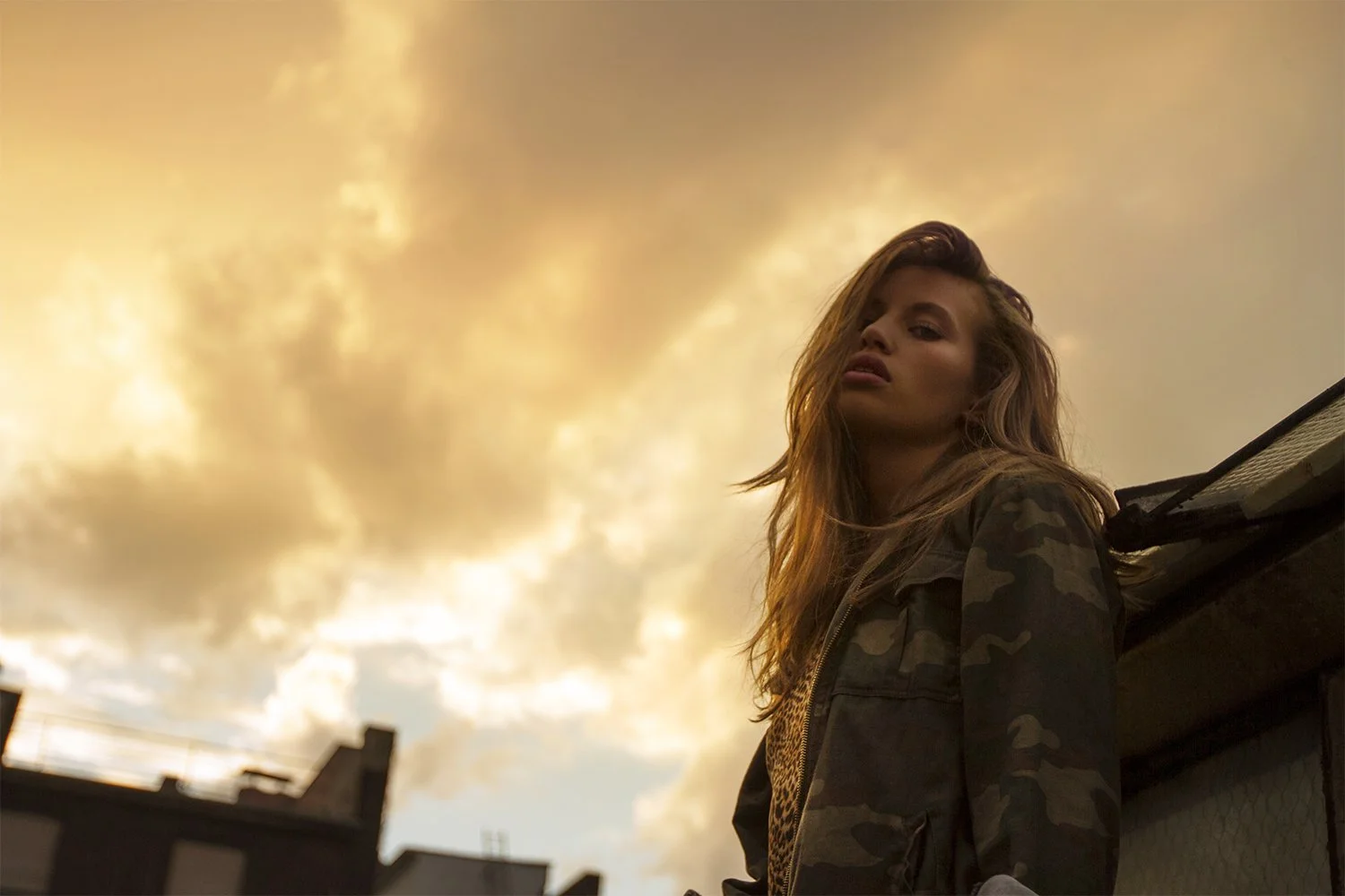 A young woman with long wavy hair stands outdoors at sunset, wearing a camouflage jacket, with a cityscape and cloudy sky in the background.