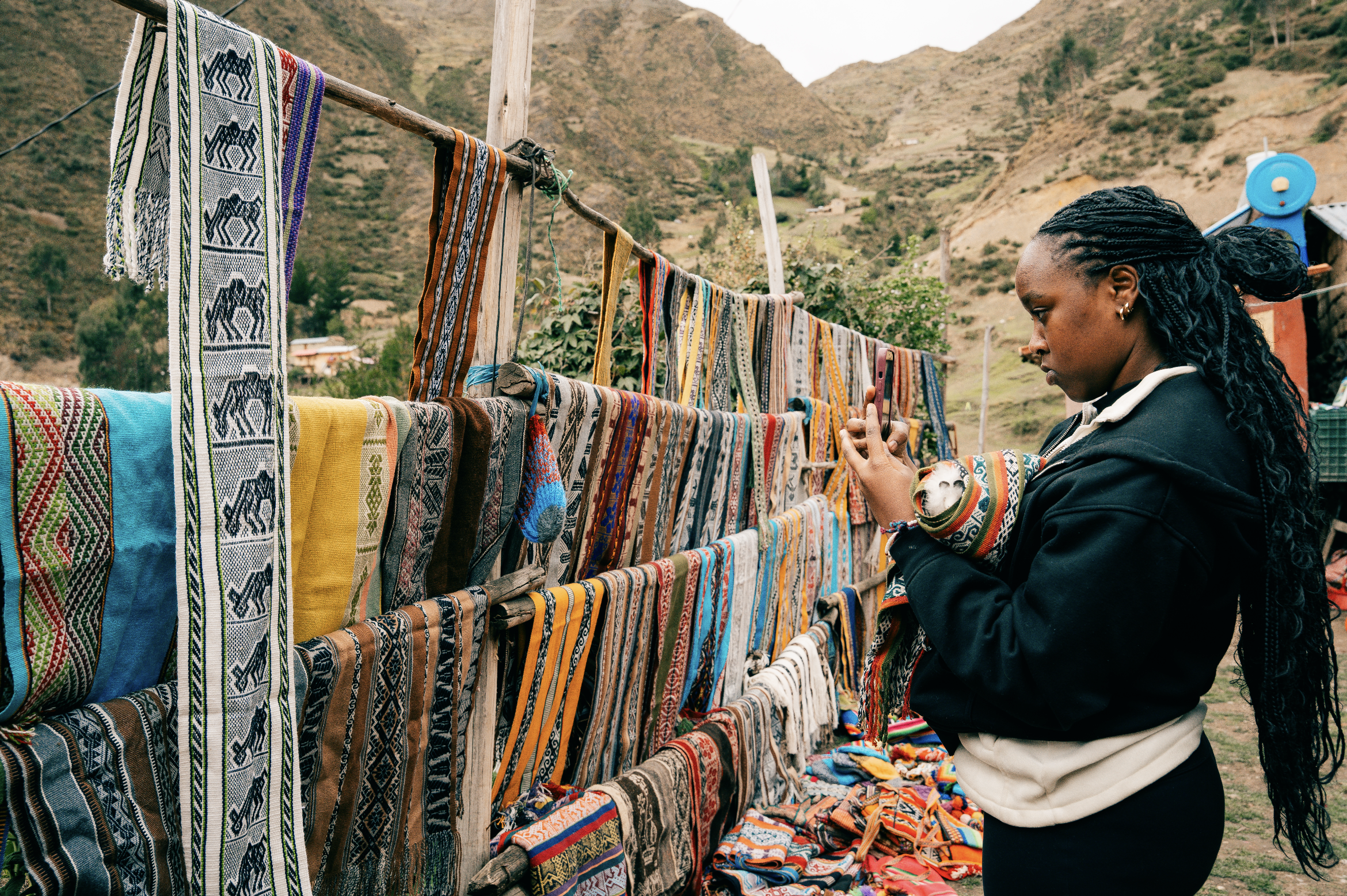 Woman at an outdoor market browsing colorful woven textiles hanging on a wooden rack, with mountains in the background.