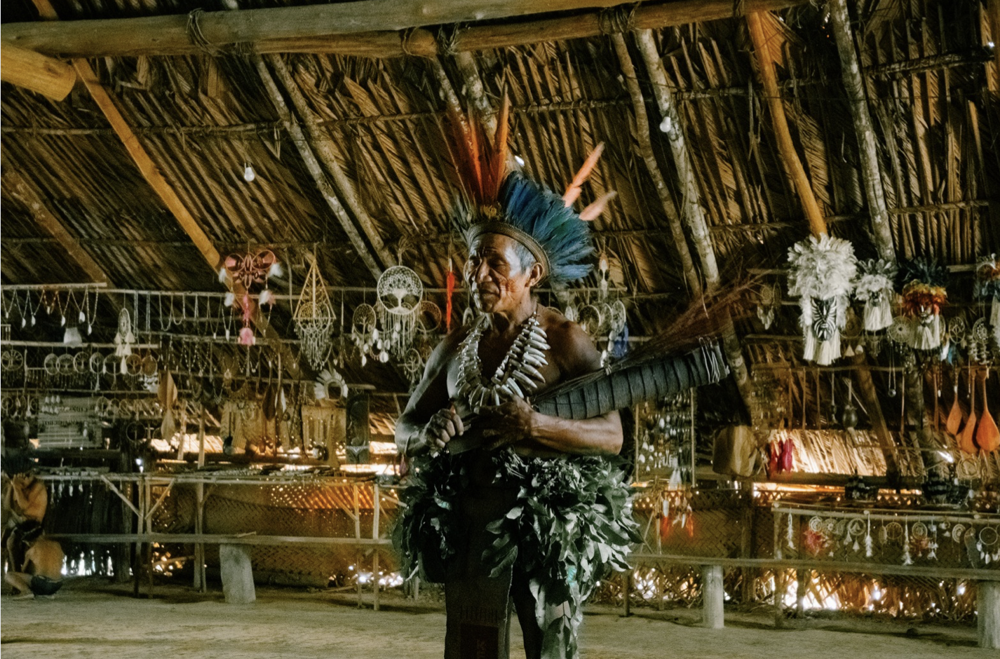 An indigenous man dressed in traditional attire with a feathered headdress and body adornments, standing inside a thatch-roofed structure decorated with various hanging ornaments and crafts.
