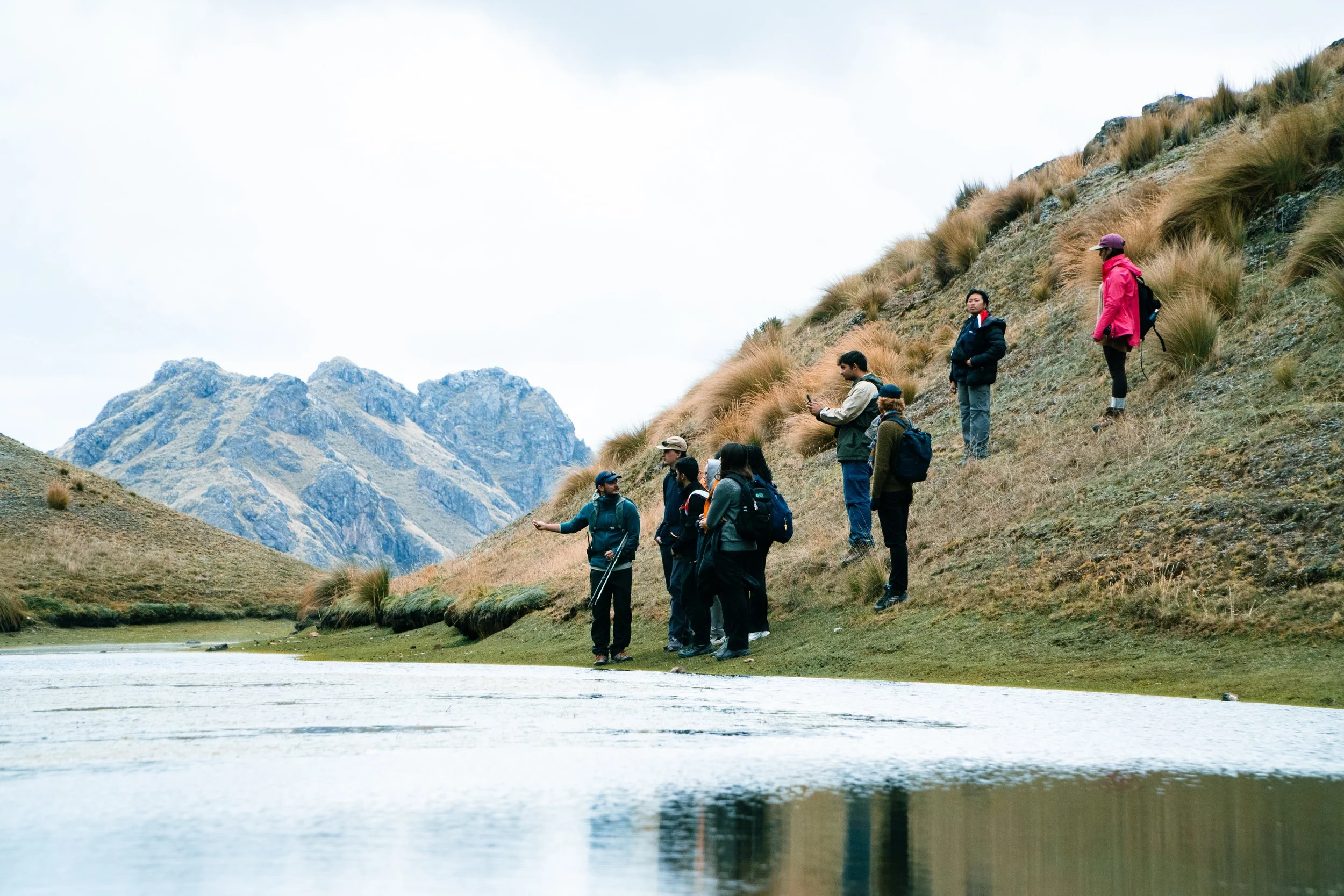 Group of people hiking near a river in a mountainous area, some talking and taking photos, with tall grasses on the hillside.