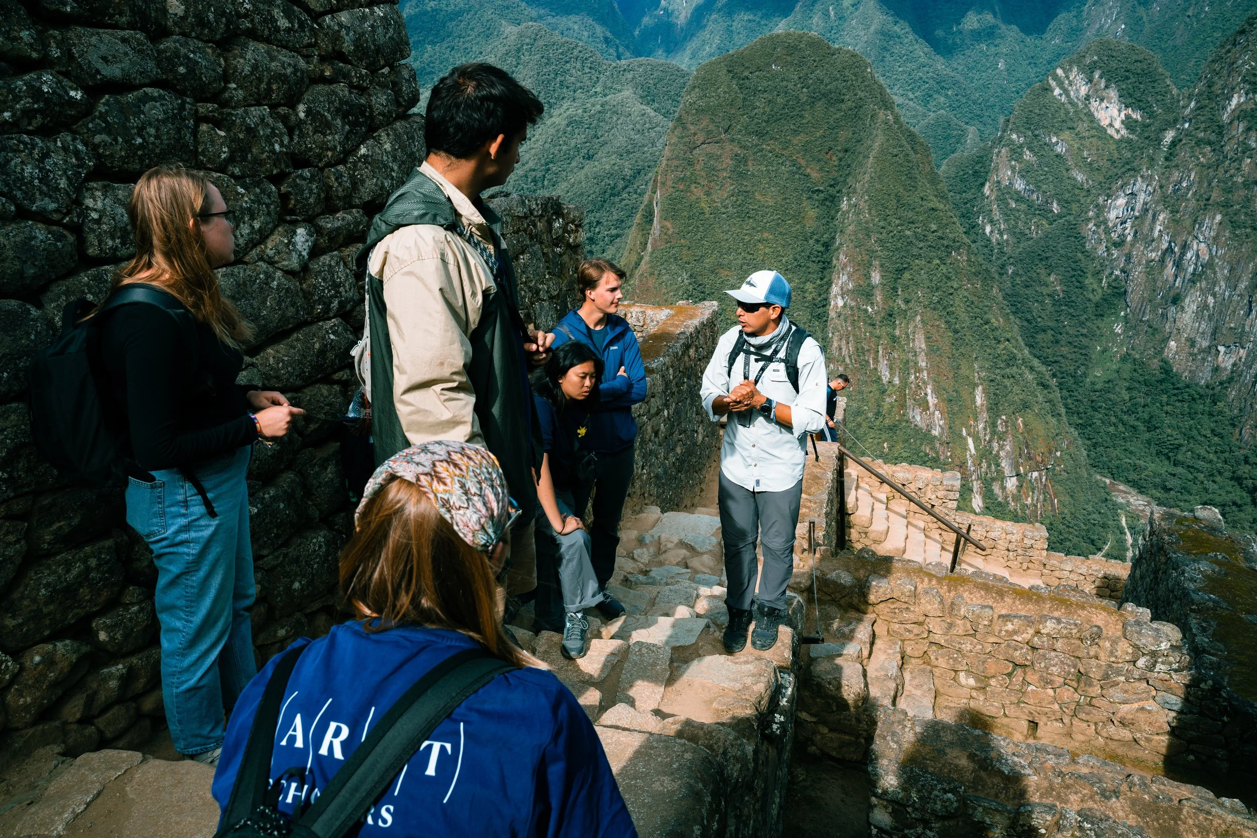 A group of tourists and a guide, all wearing outdoor clothing and backpacks, standing on stone steps at Machu Picchu, surrounded by green mountains.