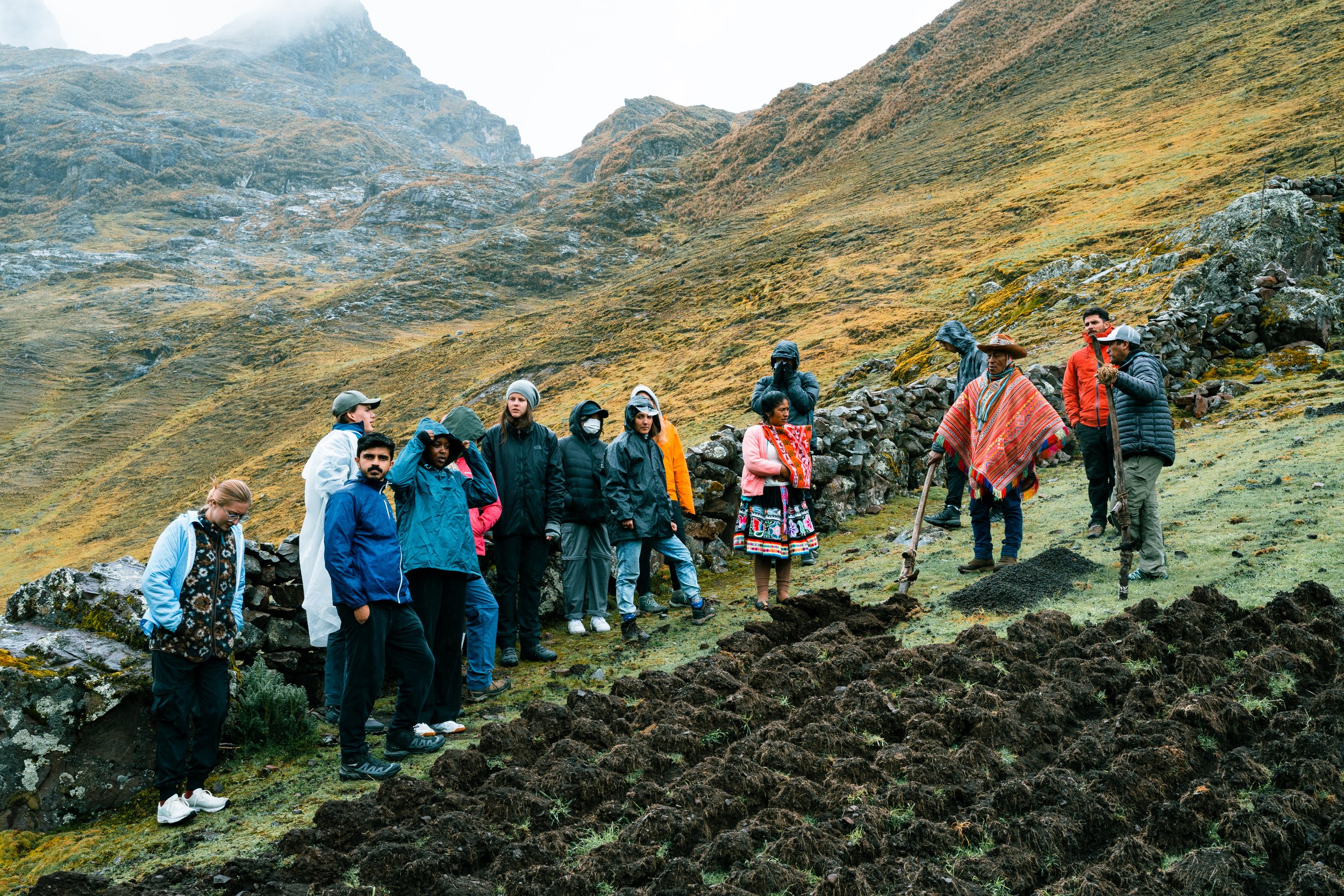 A group of tourists on a mountain hike, observing two locals in traditional clothing at a site with volcanic soil and rocky terrain.