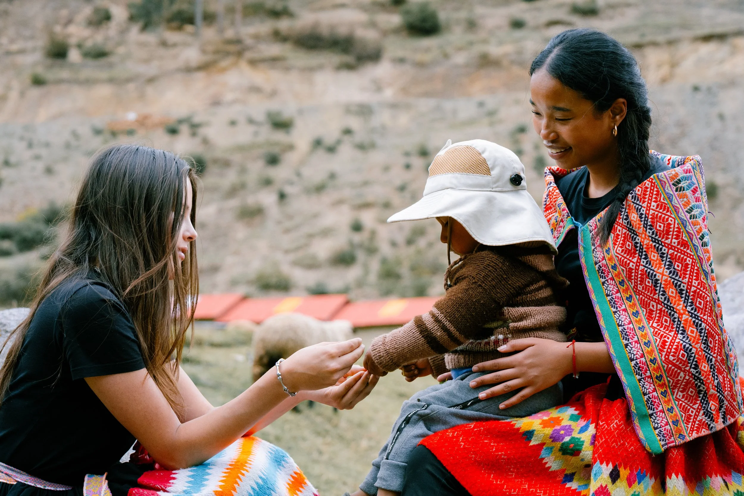 A woman with colorful traditional clothing holds a young child, who is reaching out to another young woman sitting on the ground, offering her a small object outdoors with a mountainous landscape in the background.