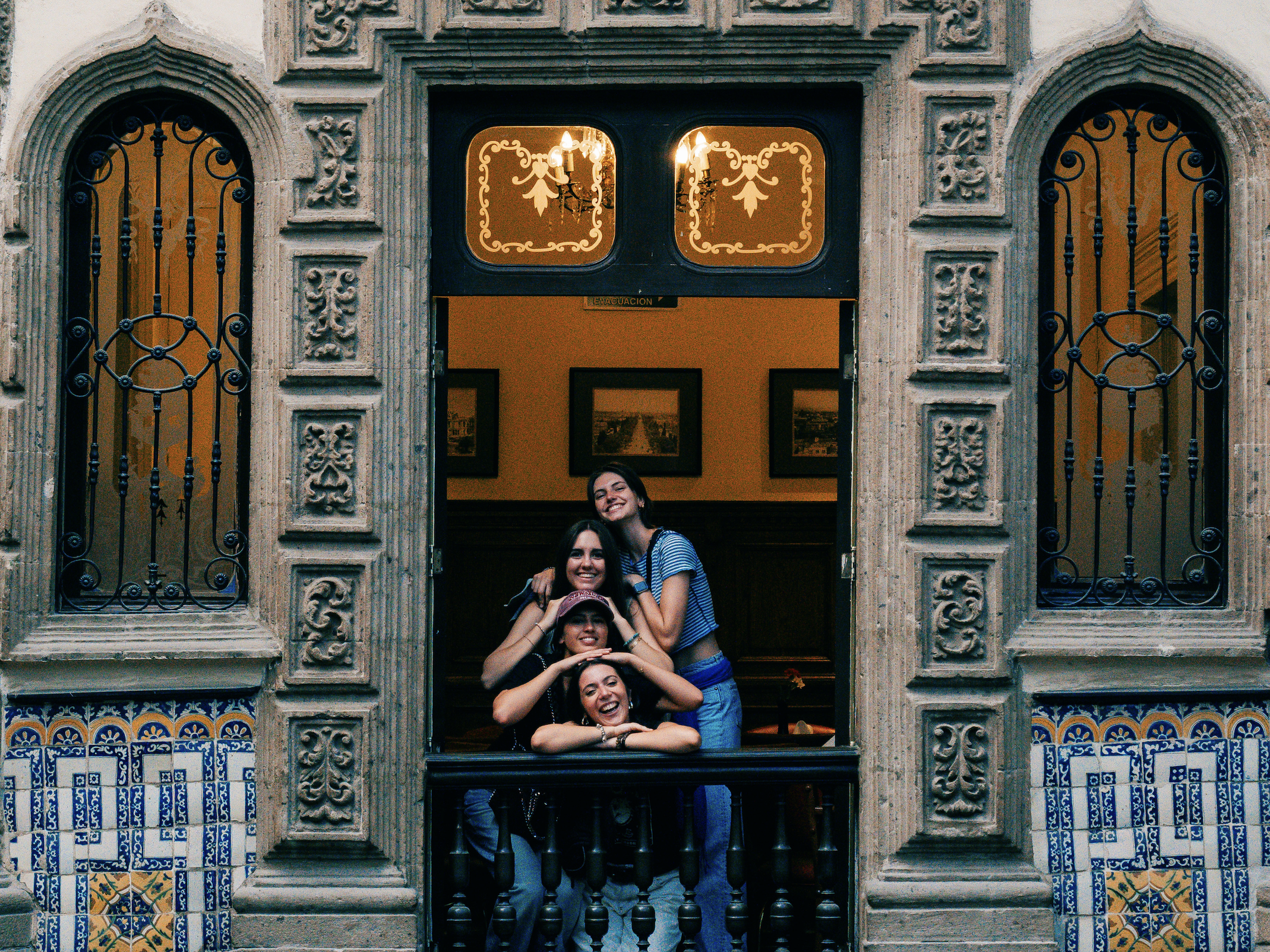 Four women posing and smiling at the open window of a historic building with ornate stonework and blue decorative tile accents.