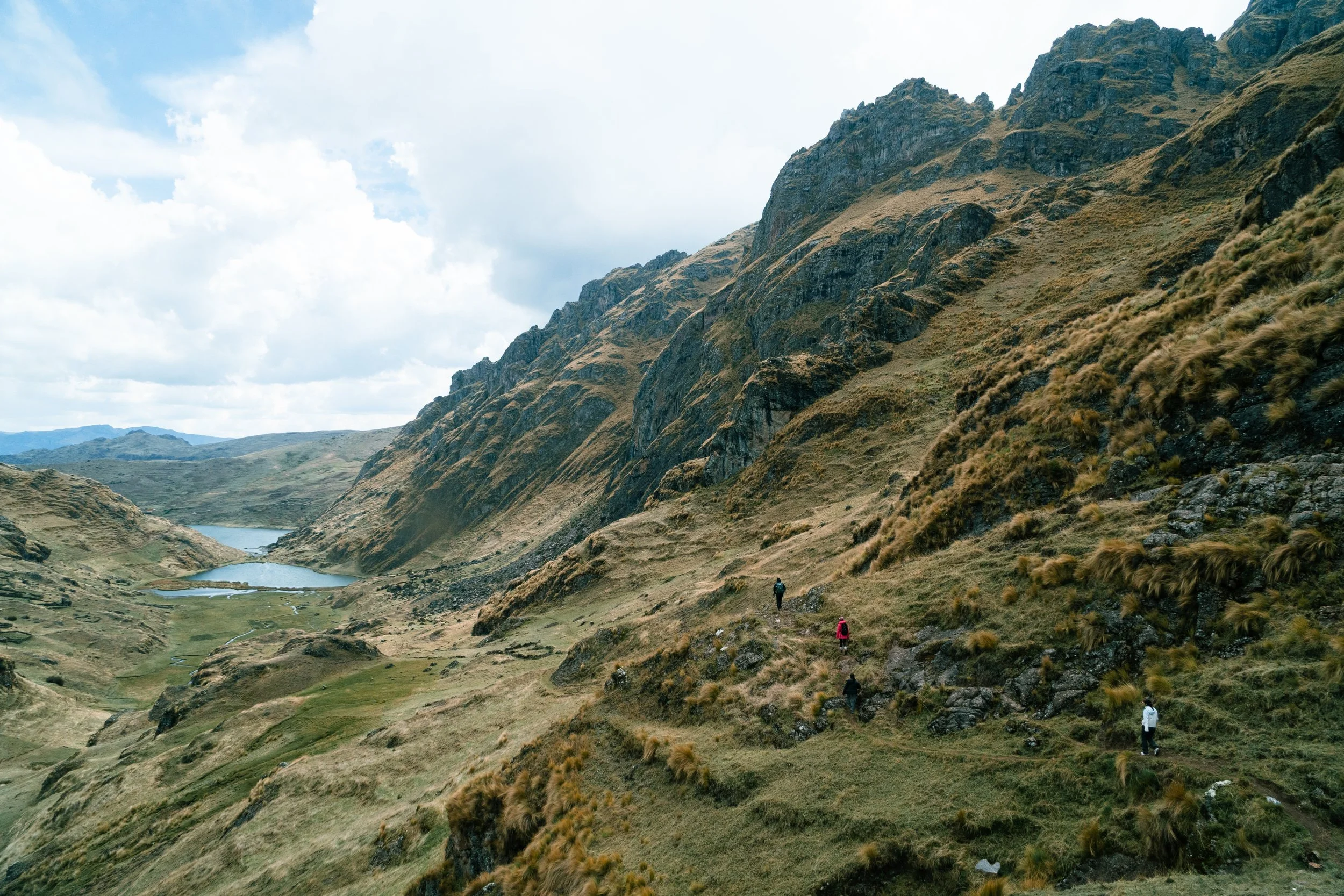 Hikers walking along a mountain trail in a valley with a lake, surrounded by steep, grassy mountain slopes under a partly cloudy sky.