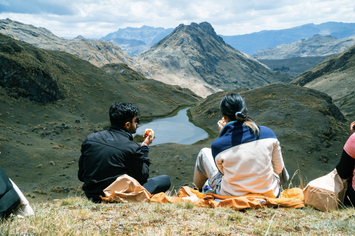 Two hikers relax on a grassy hill overlooking a mountain valley with a small lake, surrounded by rugged peaks under a partly cloudy sky.