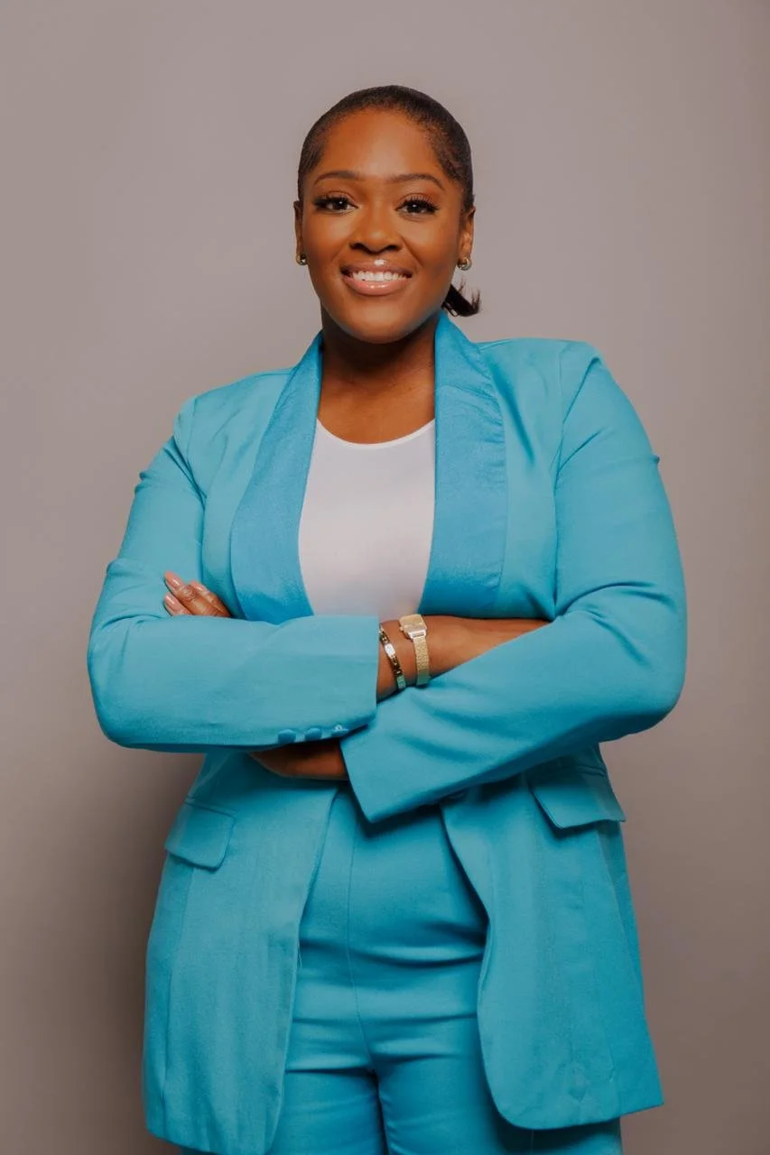 Jenny Okolo A woman with dark skin and short hair, wearing a bright blue suit over a white top, standing with arms crossed and smiling, against a plain gray background.