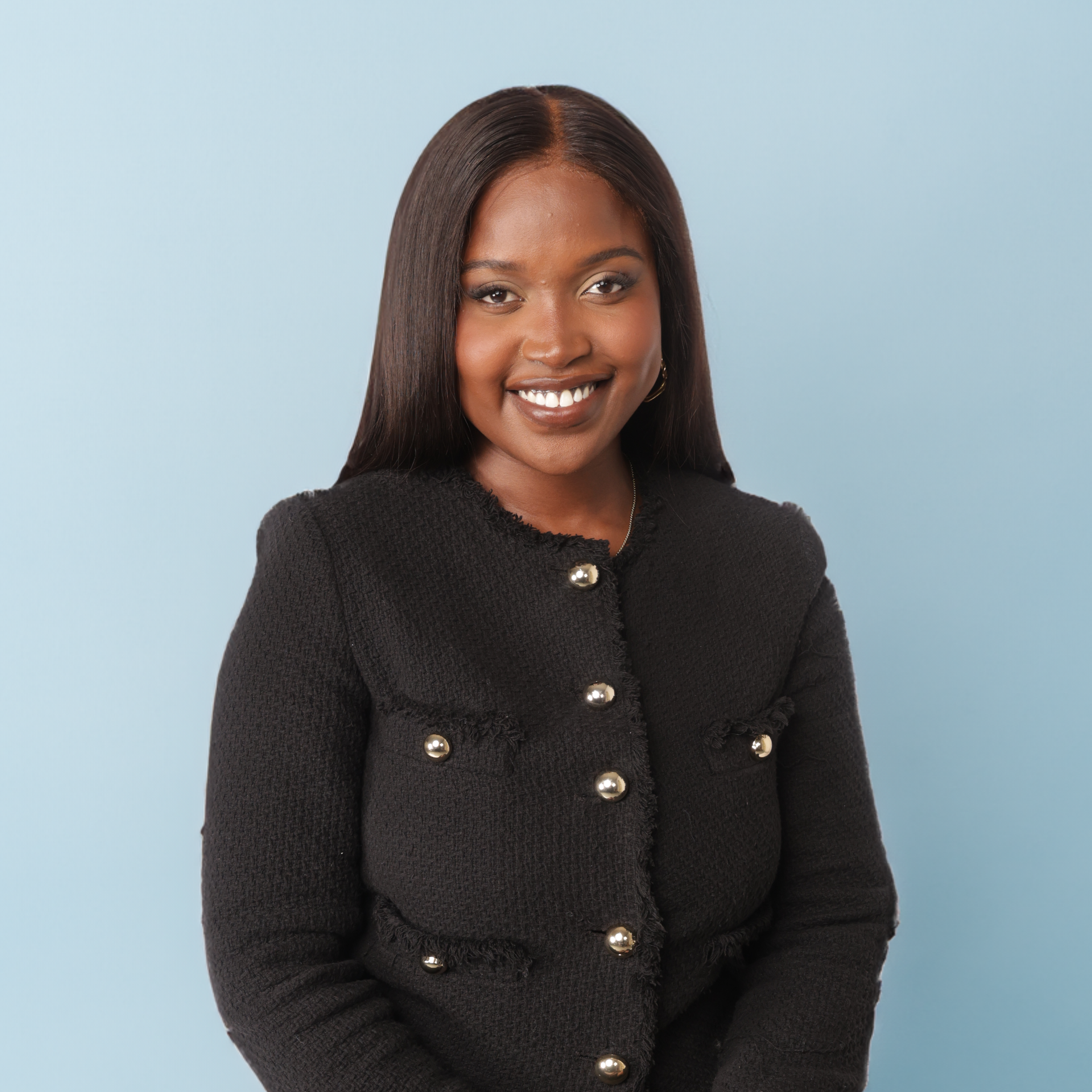 Blessing Mukosha smiling, with straight brown hair, wearing a black textured jacket with large gold buttons, standing against a light blue background.