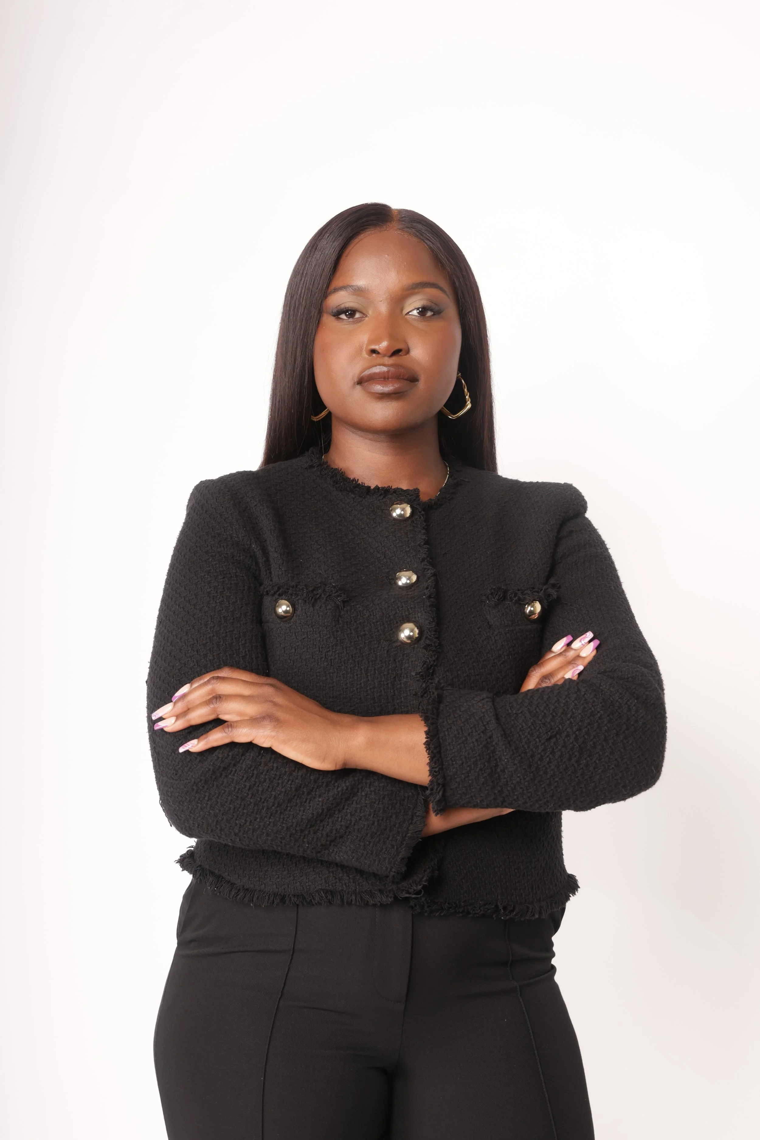 Blessing Mukosha Portrait of a confident woman with long dark hair, wearing a black textured jacket with pearl buttons and black pants, standing against a plain white background.