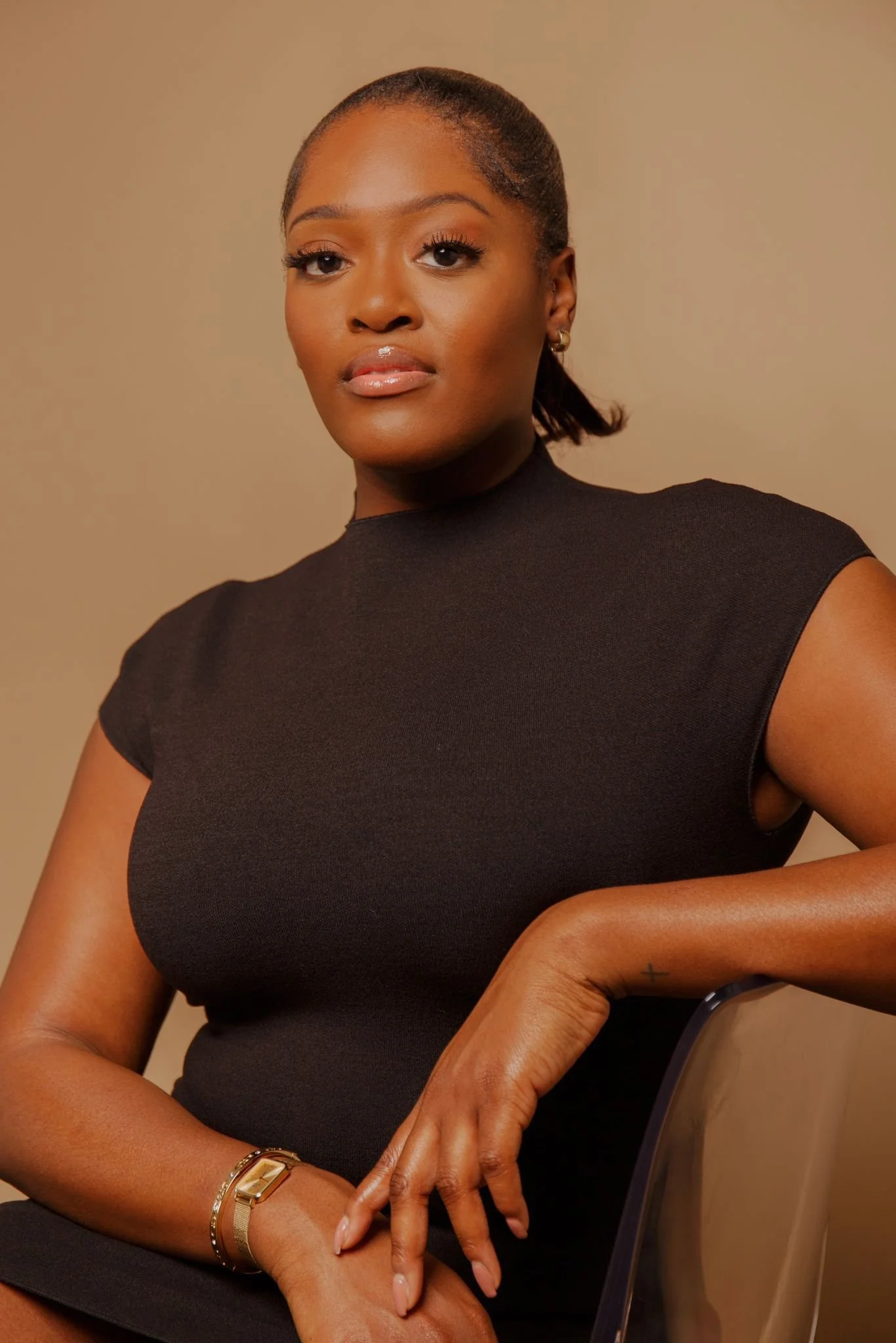 Jenny Okolo a woman with dark skin and dark hair tied back, wearing a black top, a gold bracelet, a watch, and small earrings, sitting on a beige chair against a plain beige background.
