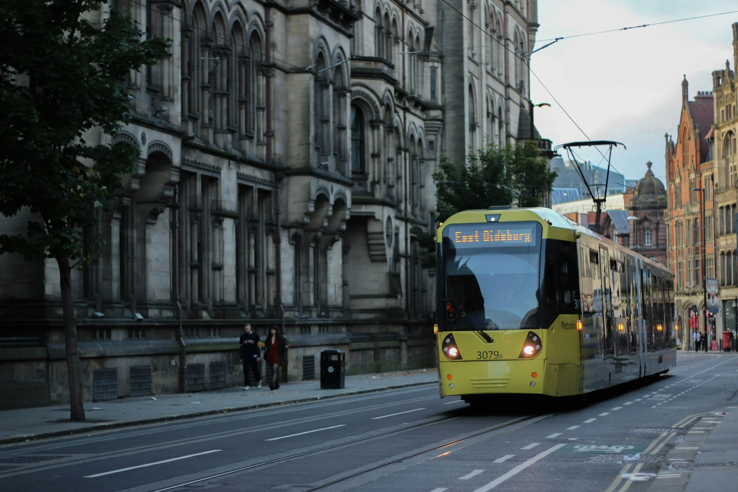 A yellow tram on a city street in front of historic stone buildings, with trees and a few pedestrians walking alongside.