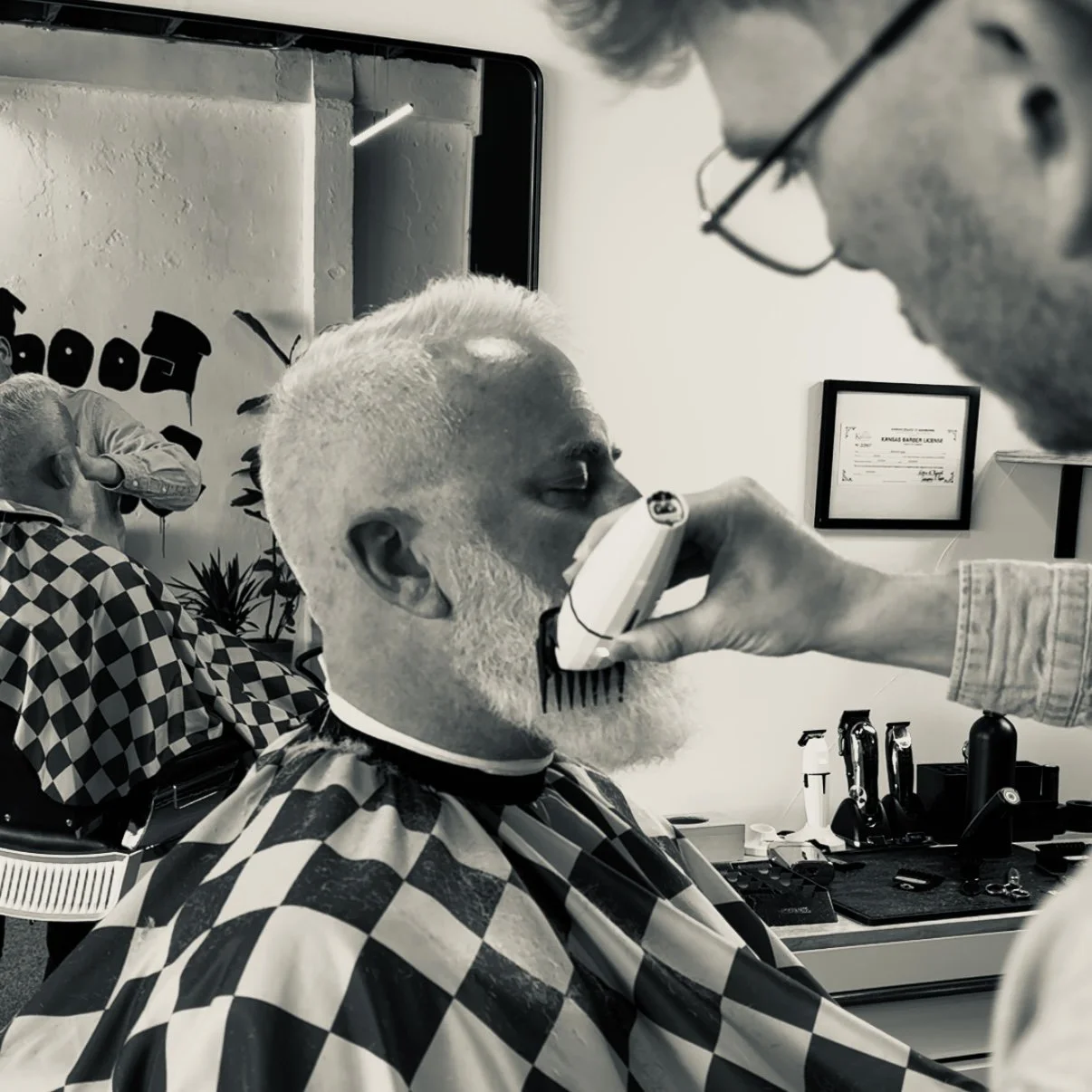 Barber trimming the beard of a customer at a barbershop, with a checkered cape around his shoulders.